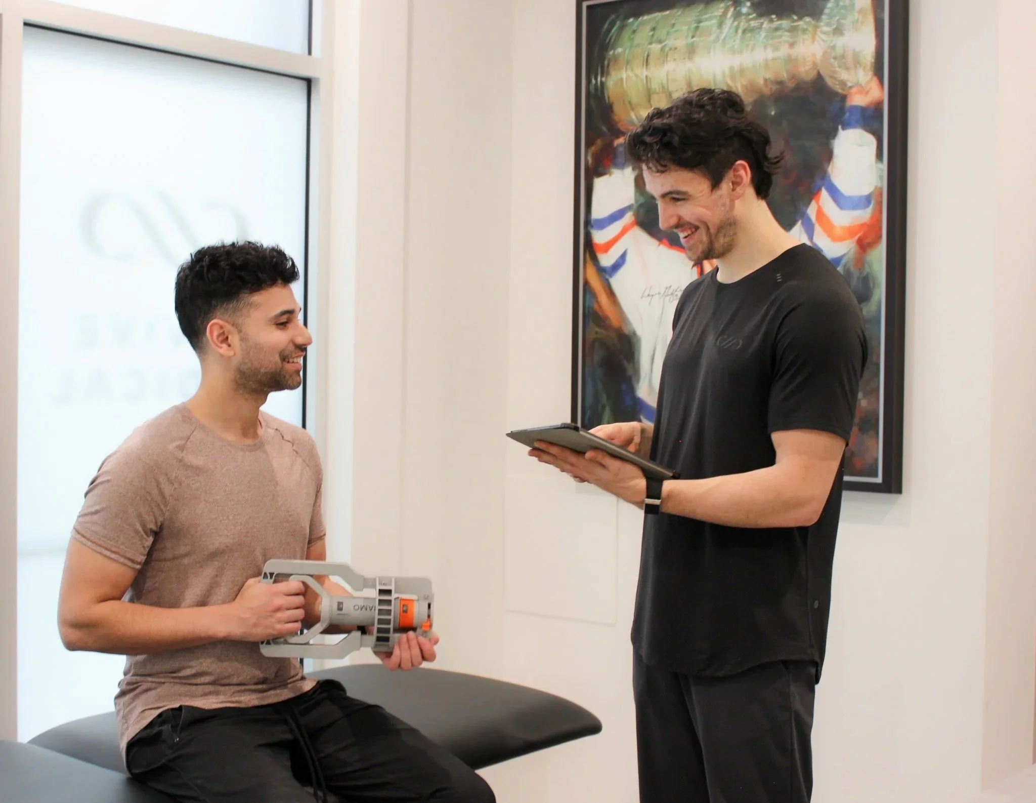 Two men are engaged in a conversation in a bright room. One man is sitting on a stool holding a small plastic device and wearing a beige t-shirt and black pants. The other man, standing, wears a black t-shirt and is holding a tablet. There is an abstract art piece on the wall behind them and large windows allowing natural light into the room.