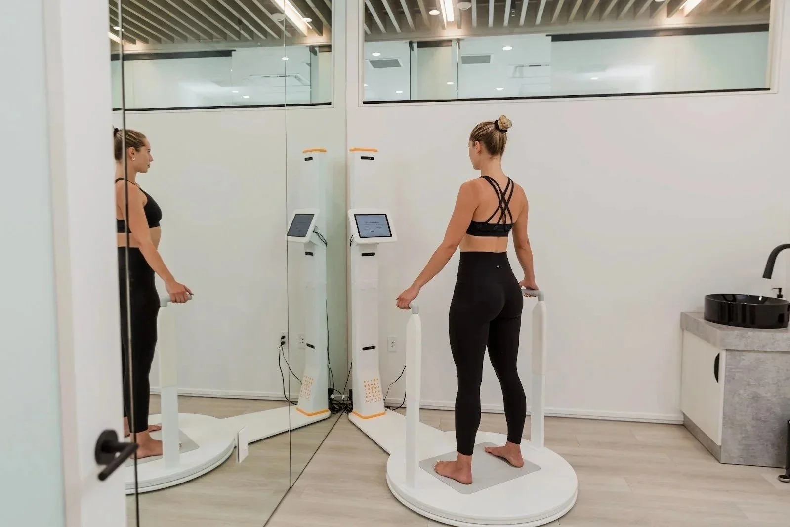A woman standing on a body composition analysis scale in a fitness studio, looking at a mirror and digital monitor, with another mirror reflecting her side view. She is dressed in black workout attire and barefoot.