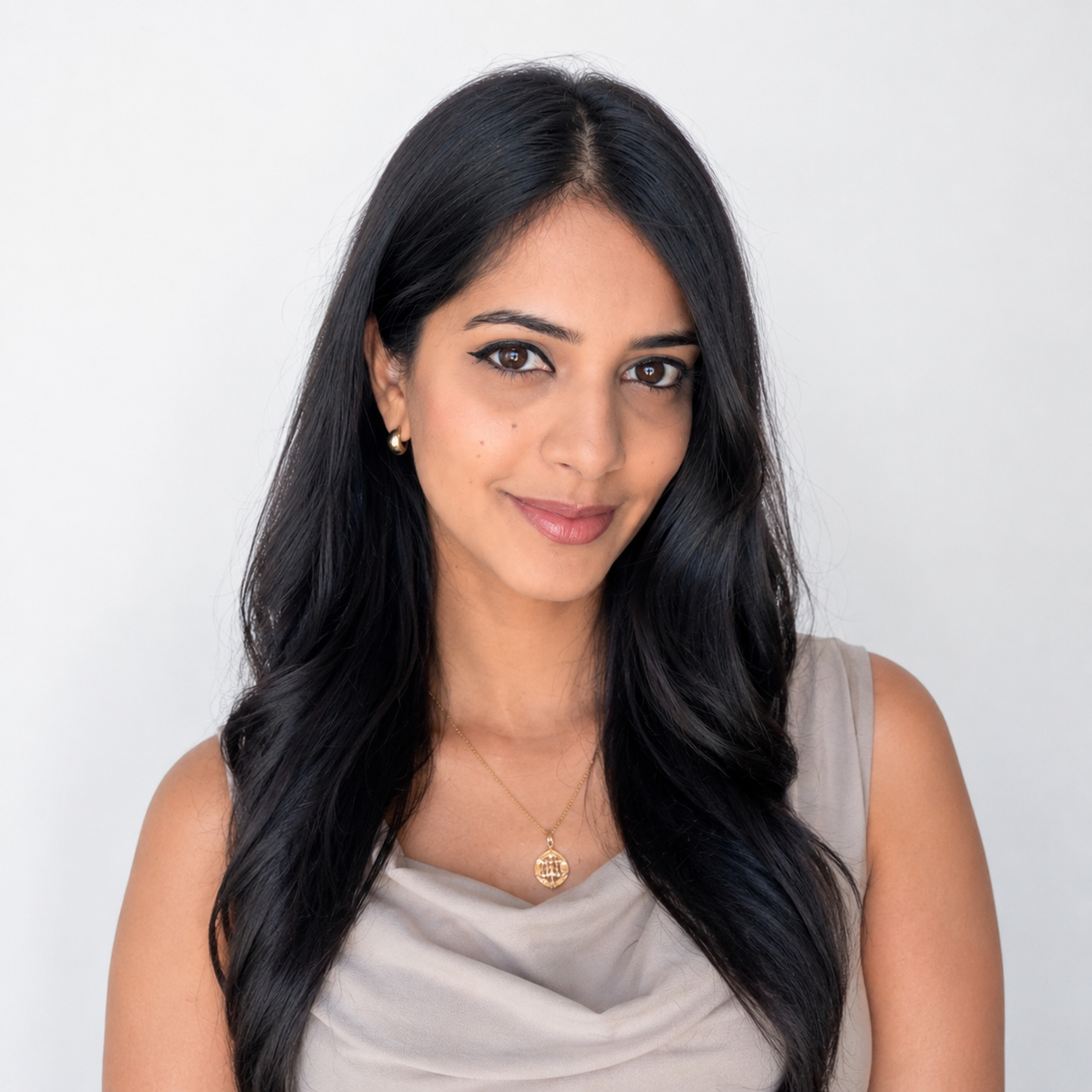 A young woman with long black hair and brown eyes, smiling, wearing a beige sleeveless top and a gold necklace with a pendant, against a plain white background.