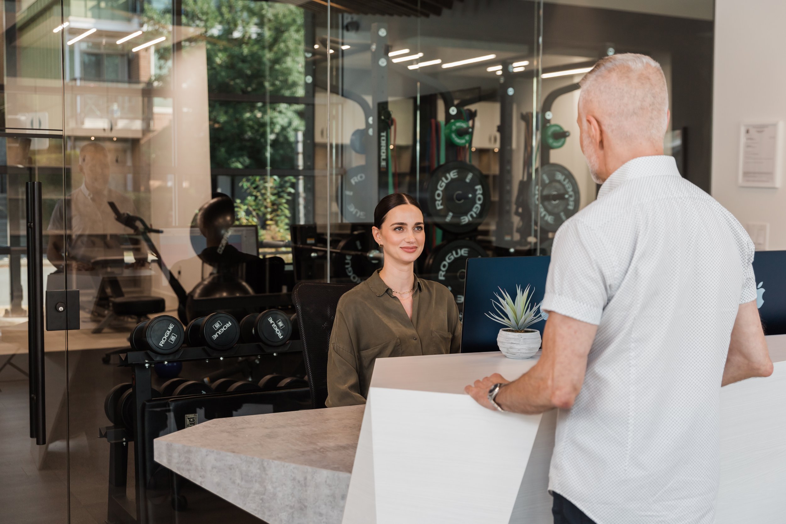 A woman sitting at a reception desk, smiling, with a man standing and talking to her inside a gym or fitness center with workout equipment visible through a glass wall.
