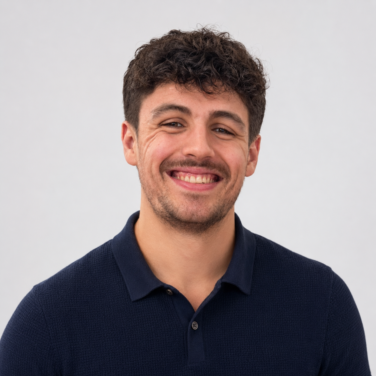 A young man with curly brown hair and light stubble smiling, wearing a navy polo shirt, standing against a plain white background.