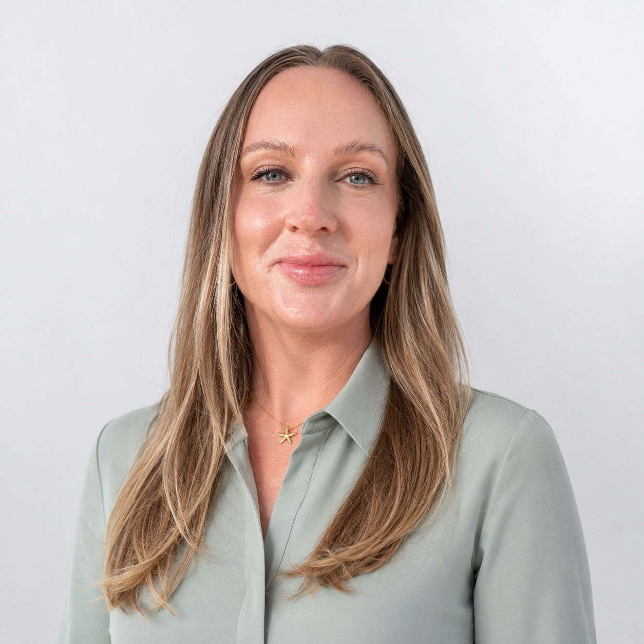 A woman with light brown, wavy hair and blue eyes, wearing a light green button-up shirt and a gold starfish necklace, smiling gently at the camera against a plain white background.