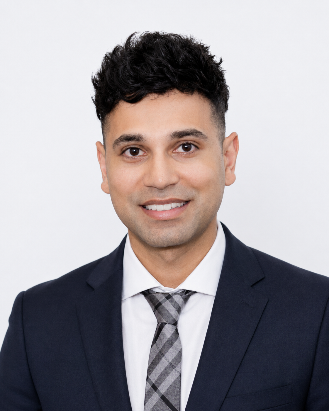 A professional headshot of a young man with dark, curly hair wearing a navy suit, white shirt, and patterned gray tie, smiling against a plain white background.