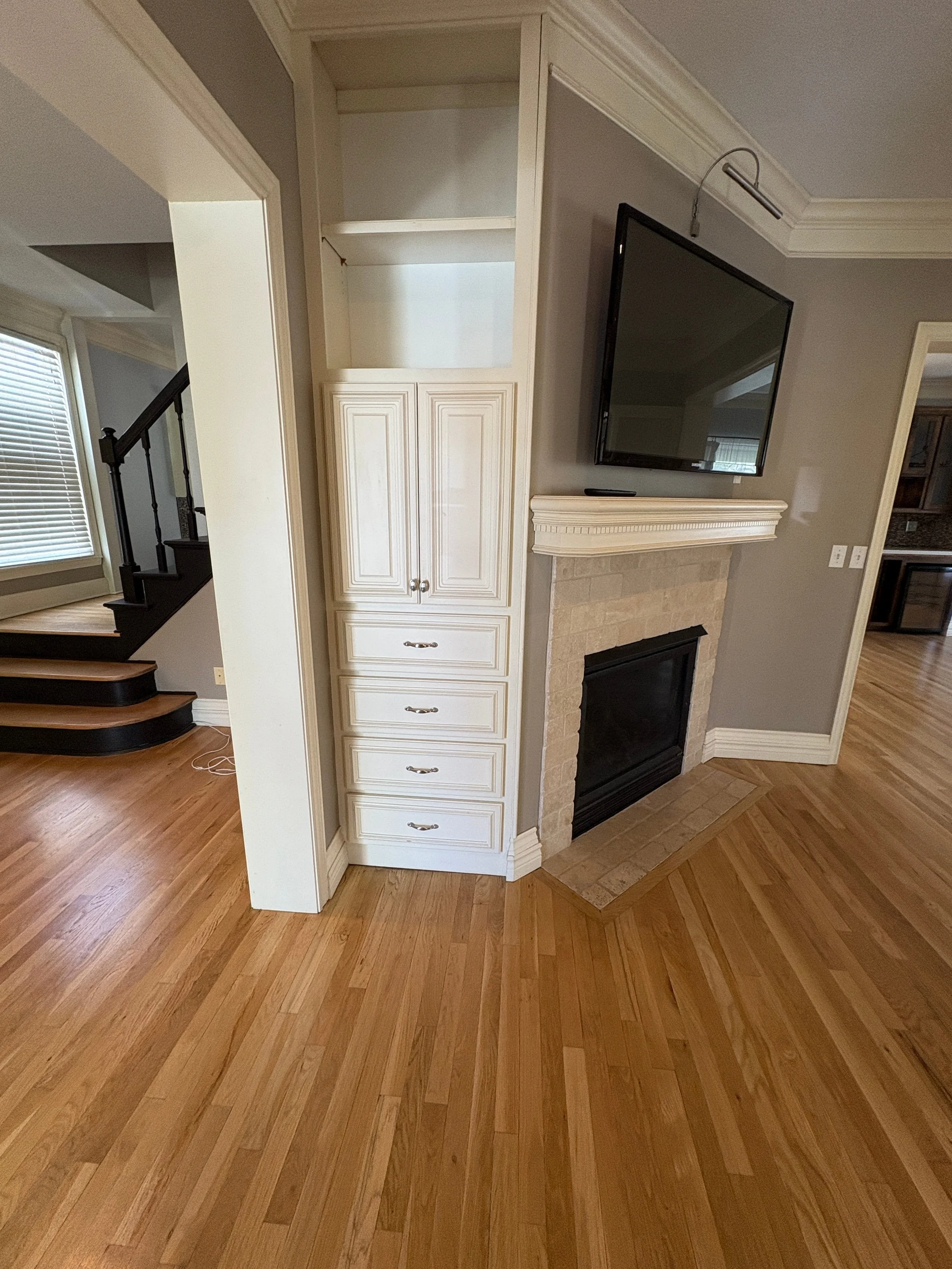 View of a newly painted beige interior living space with wood flooring with painted white drawers and white trim. A fireplace with a TV above it is on the right and a staircase is to the left.