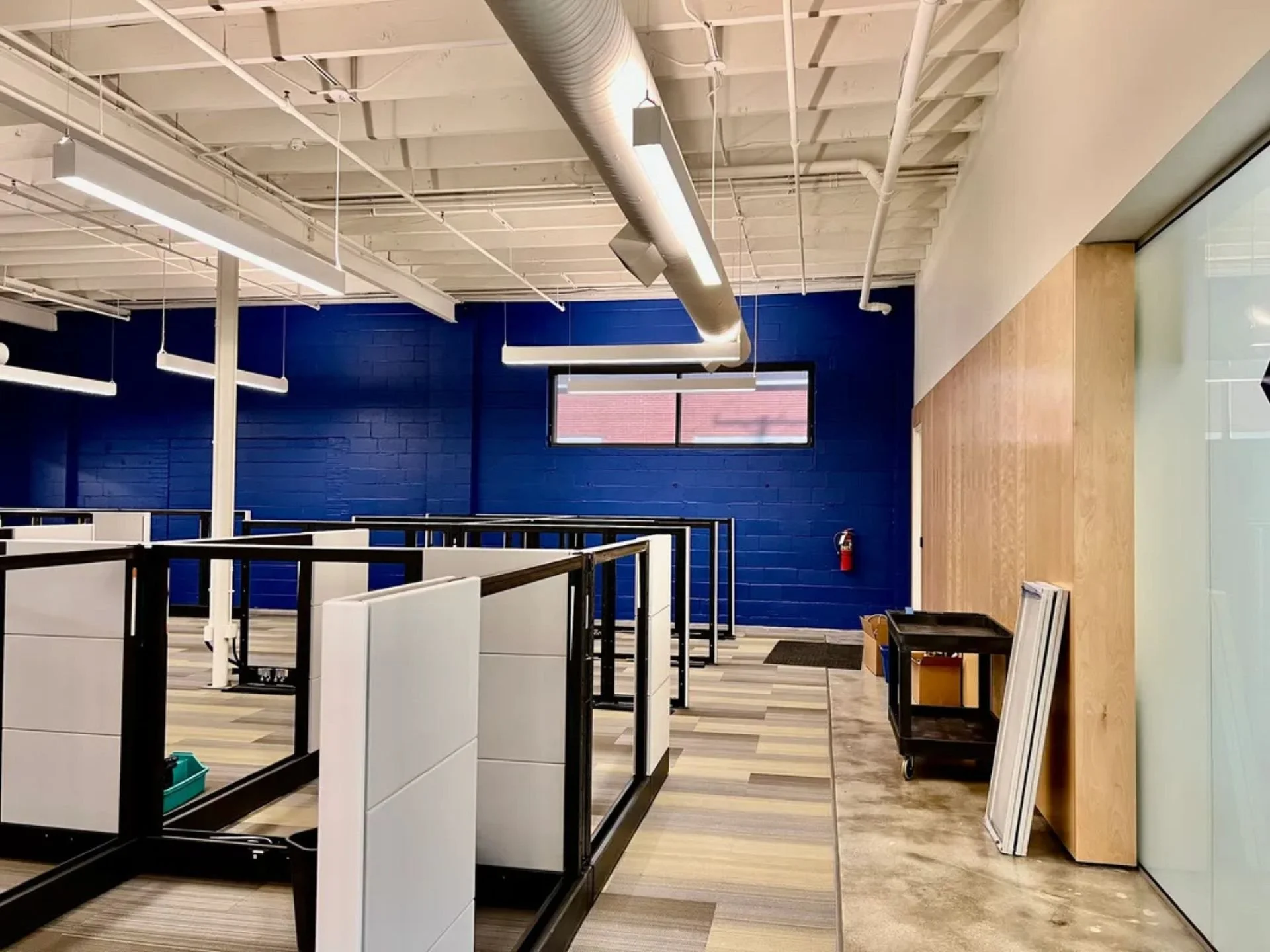 An interior space with cubicle dividers and a newly painted blue brick wall in the background. There are ceiling lights and an exposed duct along with a window showing a red roof outside. The space appears to be under construction or setup.