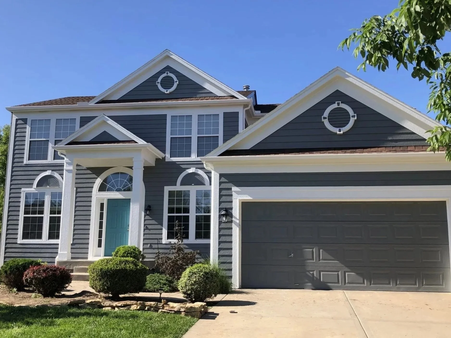 Front view of a two-story house in Kansas City with dark gray siding, white trim, a teal front door, multiple large windows, a bush-lined walkway, and a two-car garage under a clear blue sky.