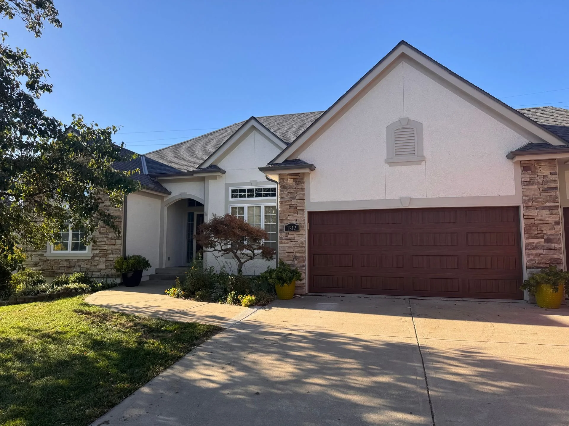 A two-story house in Overland Park, Kansas with a brown garage door, newly painted beige stucco exterior walls, a front yard with bushes and a tree.