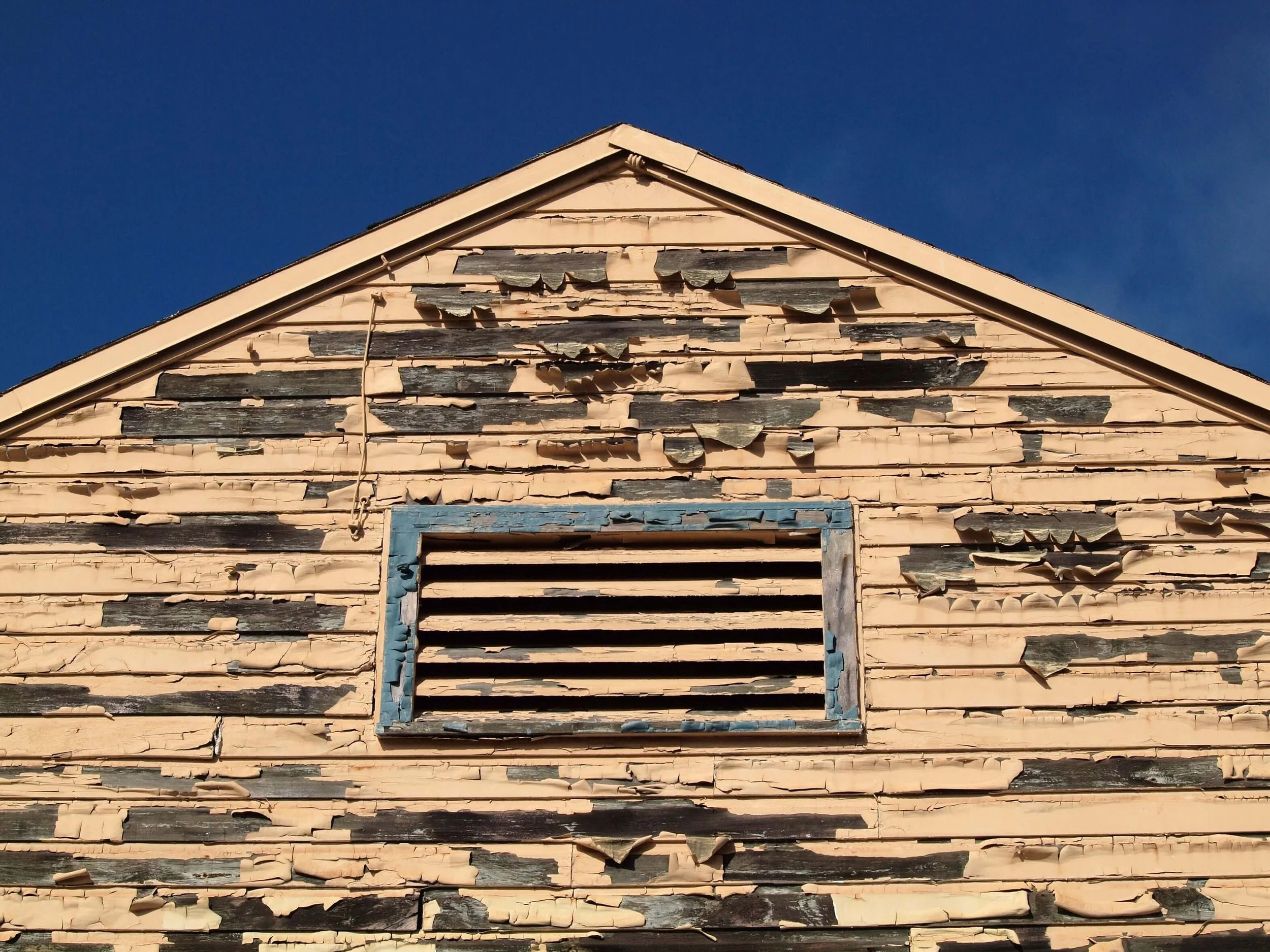 A view of the top of the side of an old house with peeling yellow paint on wooden siding showing wood rot in need of repair.
