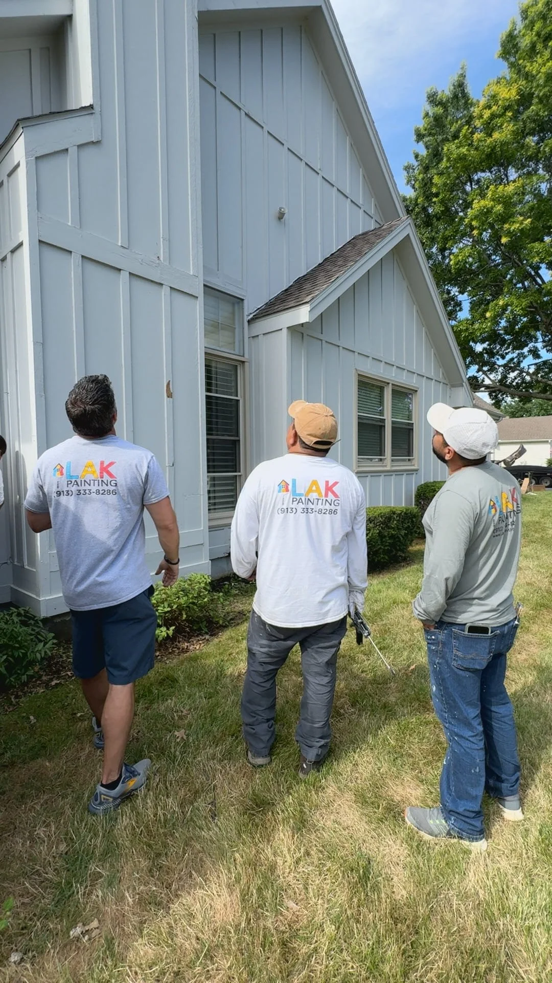 Three workers in LAK Painting uniforms inspecting the exterior of a white house with siding and windows, standing on a grassy yard on a sunny day.