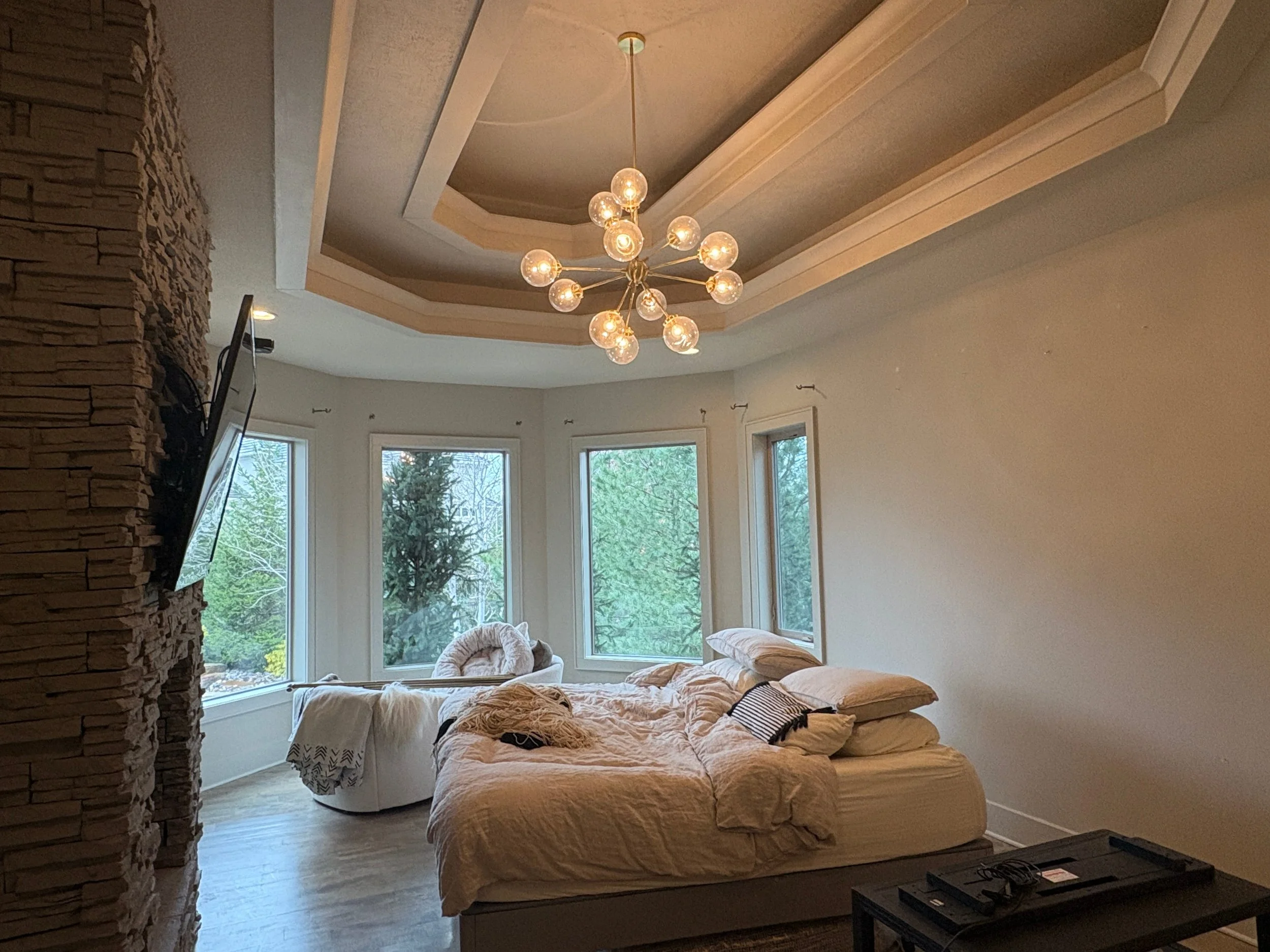 A newly painted beige bedroom with large windows showing trees outside and a stone wall on the left side. The ceiling has a beige oval inset molding feature and a modern chandelier with multiple light bulbs hanging from the ceiling
