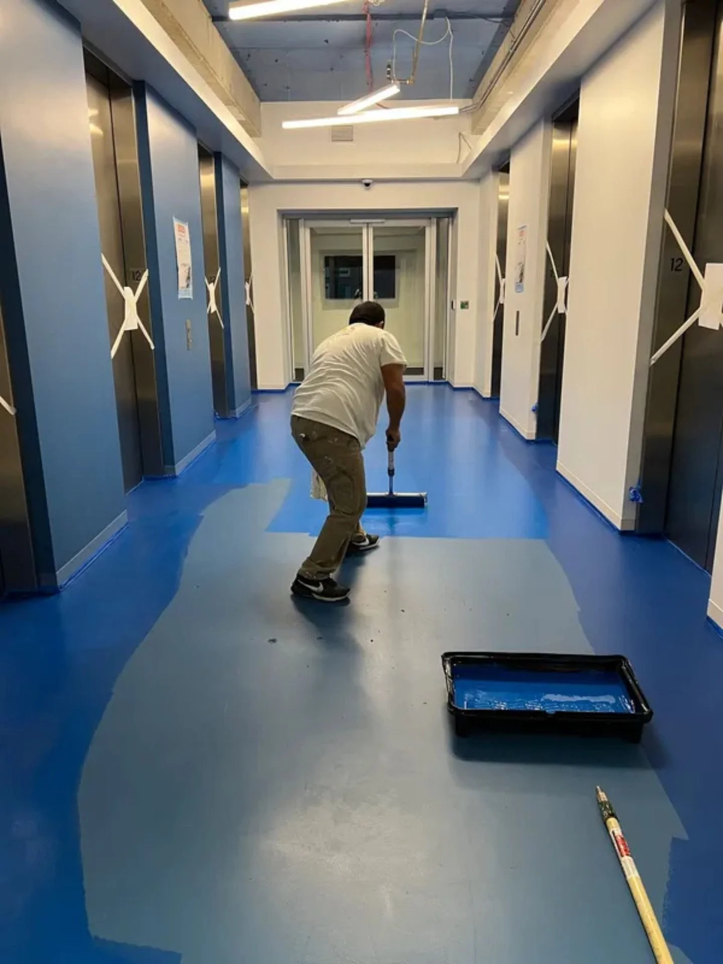 An LAK Painting commercial painter in a Kansas City office lobby between closed elevators painting the floor a bright blue, holding a long painting tool next to a paint basin.