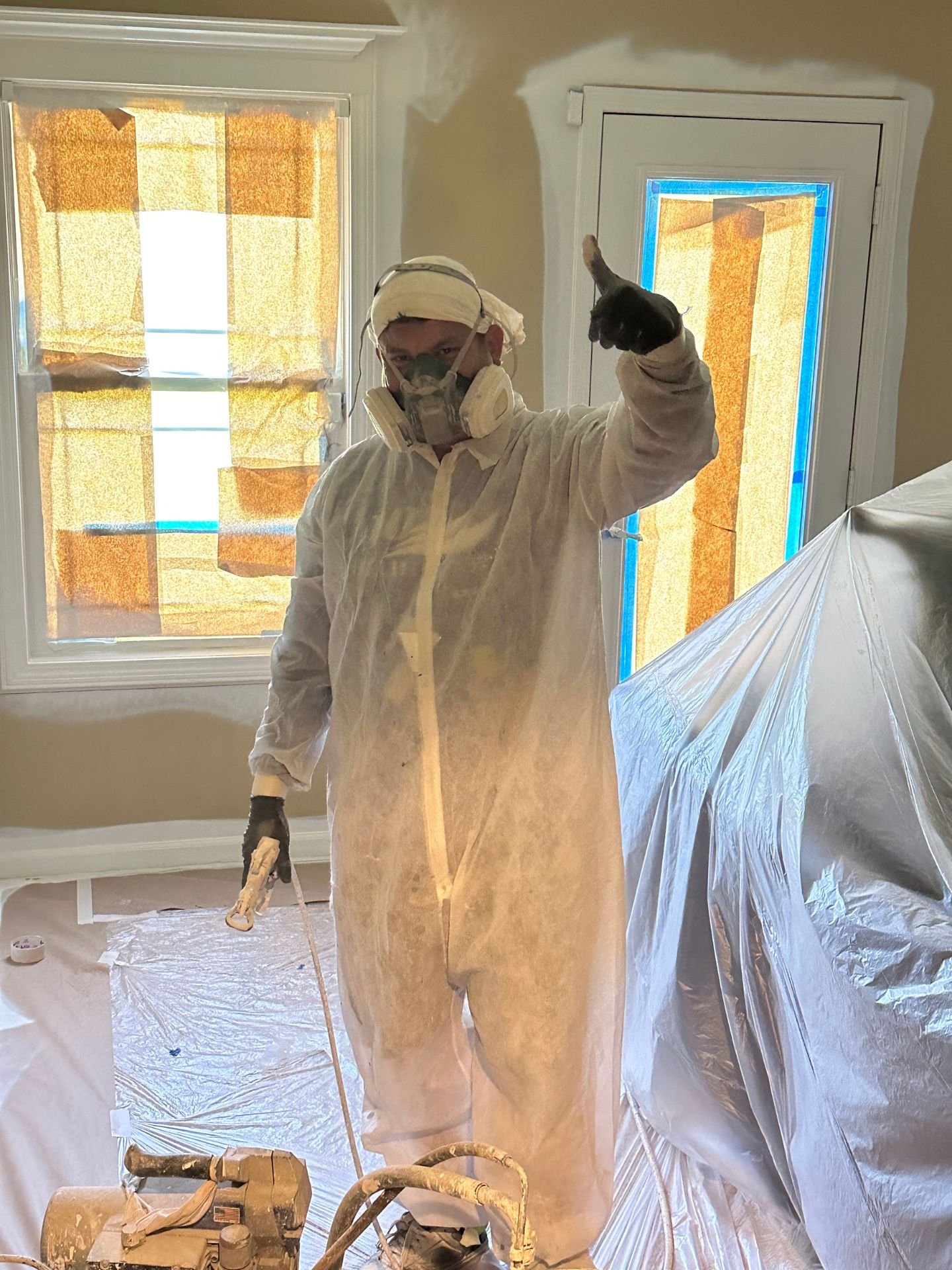 An LAK Painting employee wearing plastic protective gear holding a thumbs-up standing in a room being painted, with plastic sheeting on the floor and window.