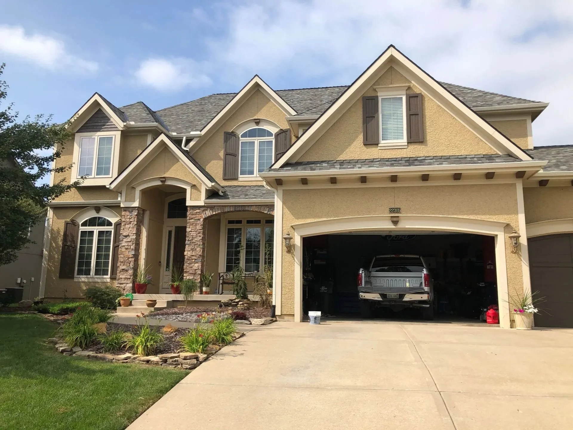 A nice two-story yellow-beige house in Overland Park, Kansas with a garage open to a driveway. The house has various windows, a small front porch, and a landscaped yard with plants and flowers.