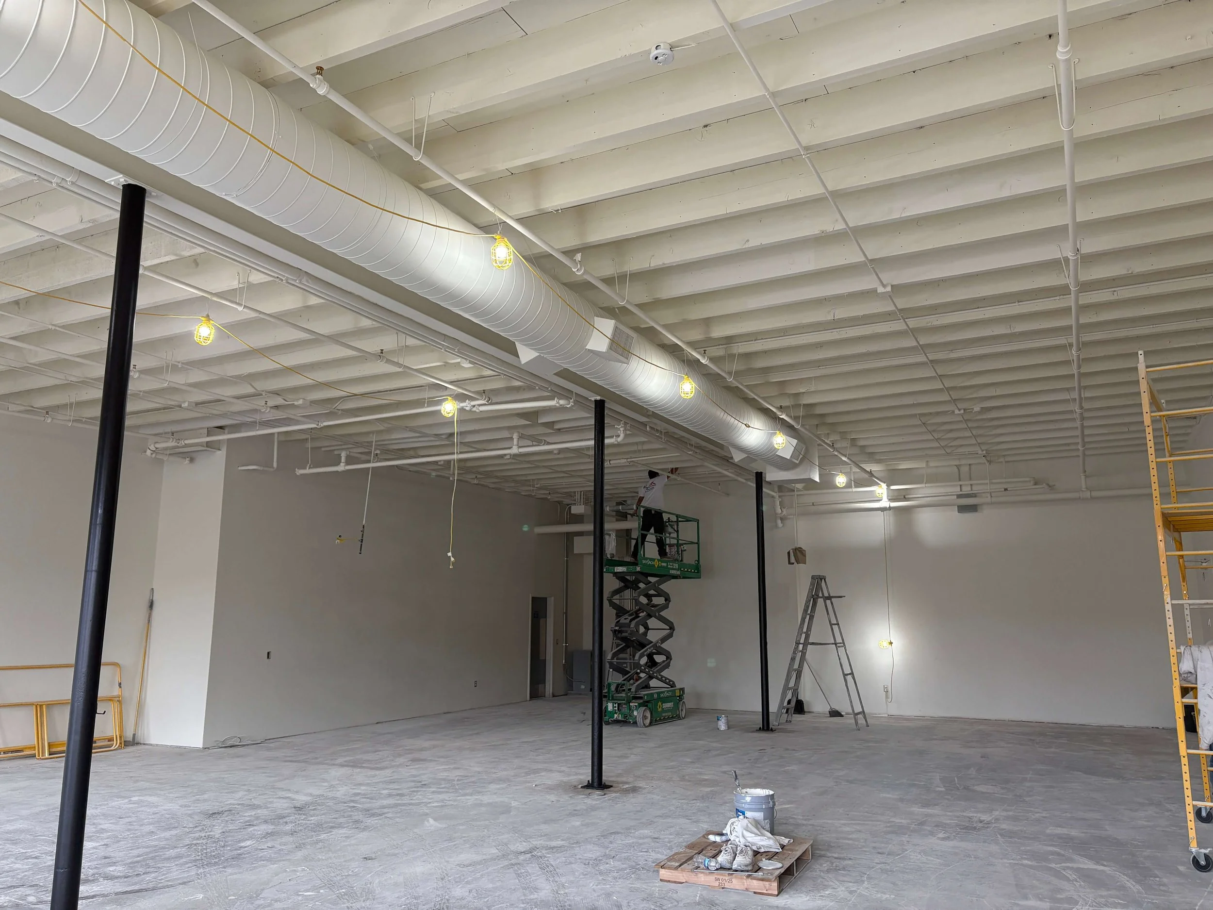 ACI Gym newly painted white by LAK Painting in Kansas while under construction with high ceilings, exposed ductwork, and scaffolding. The walls are white and there is a concrete floor with three black structural posts.