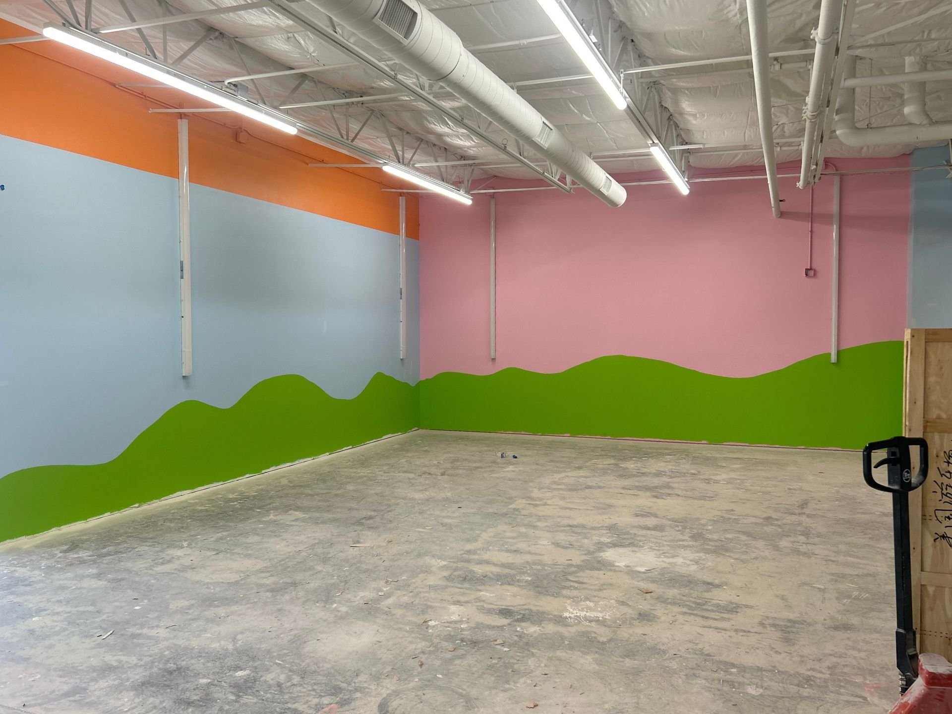 Interior of Kids Empire in Kansas with newly painted concrete walls, bare concrete floors and industrial lighting. The farthest wall is pink and the wall on the left is light blue with a green hill painted motif going across both walls.