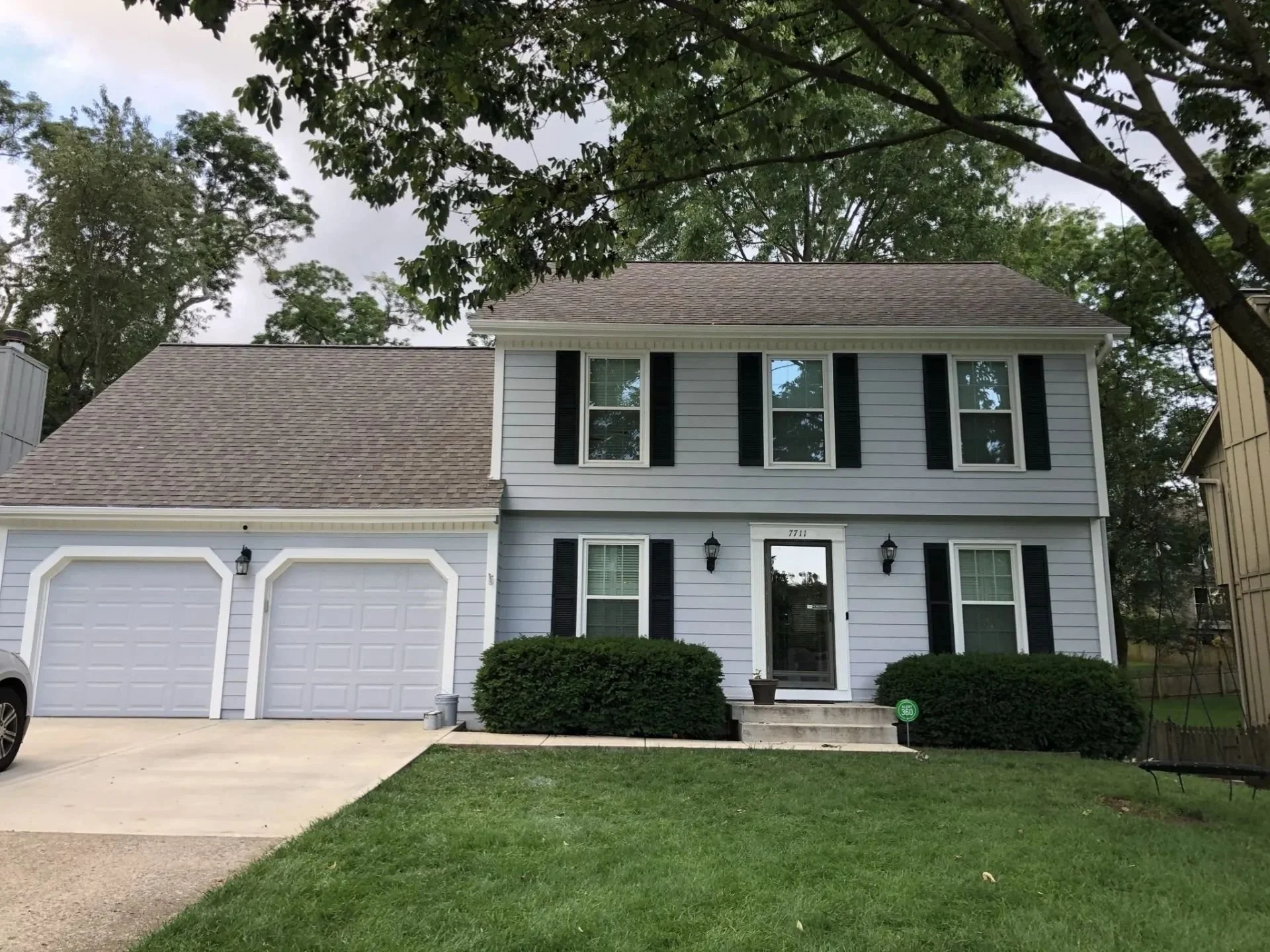 A colonial style two-story house in Overland Park, Kansas with light blue-gray siding, black shutters, and a shingled roof. It has a two car garage, a green lawn, and bushes at the front of the house with large oak trees behind the house.
