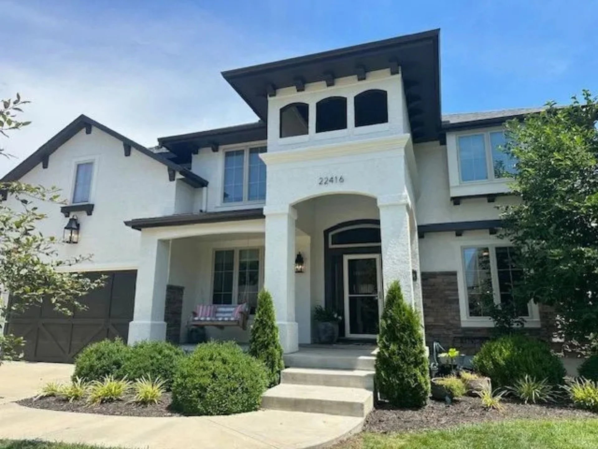 Front view of an impressive modern two-story house in Shawnee, Kansas with a newly painted white exterior, dark trim, and an arched covered entryway. Landscaped with bushes with a concrete walkway leading to the front door.