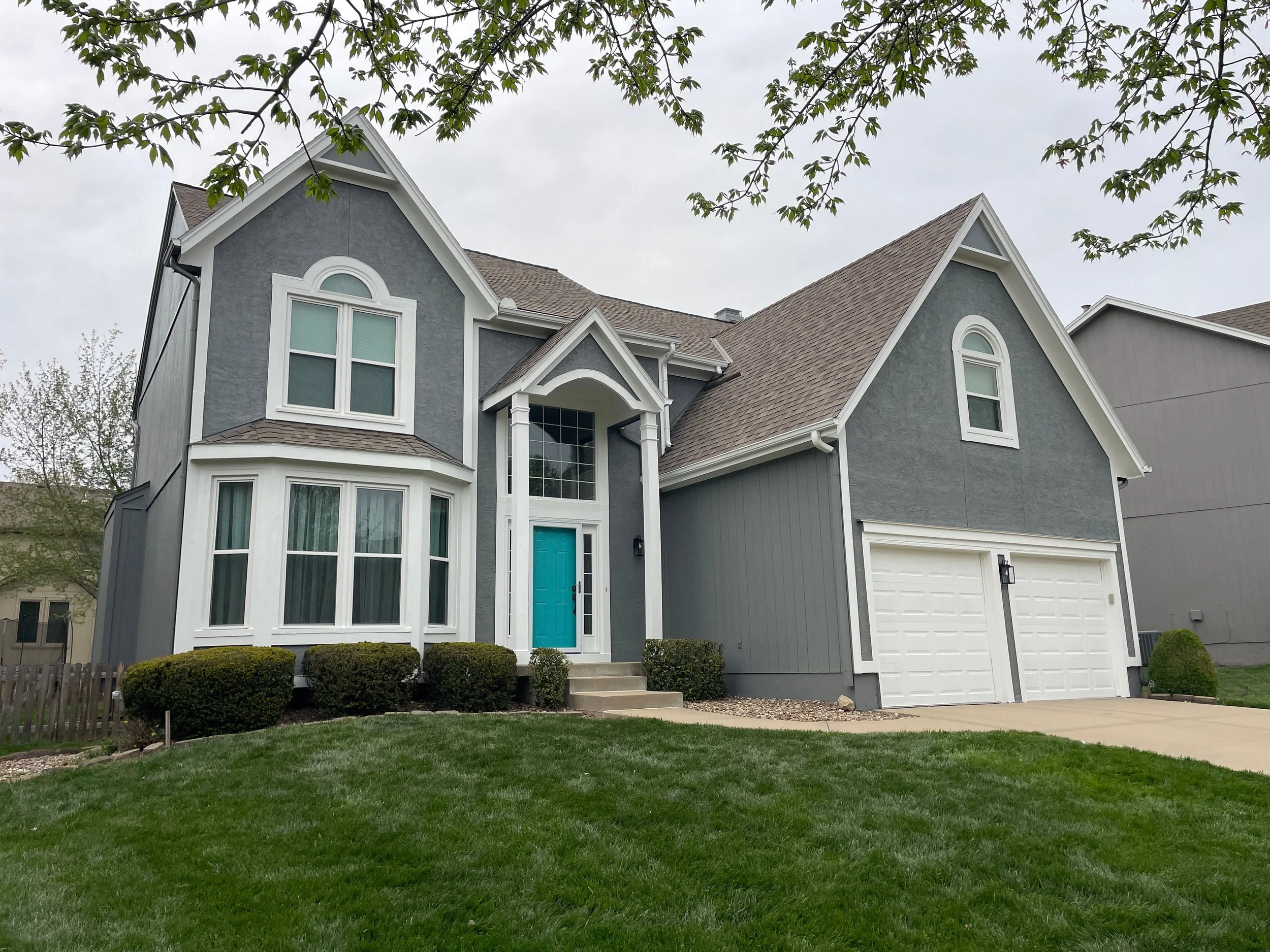 A large freshly painted gray house in Lenexa, Kansas with white trim and garage doors, a teal front door, multiple gables, and white window frames. There is a manicured lawn with bushes in front and a sidewalk leading to the entrance.