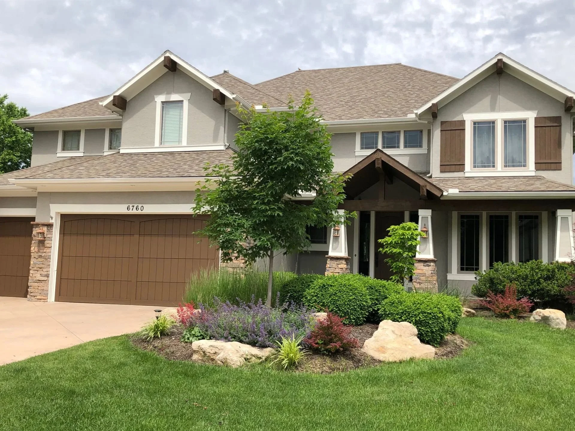 A two-story house in Overland Park, Kansas with a brown garage door, newly painted beige stucco exterior walls, a front yard with bushes and a tree.
