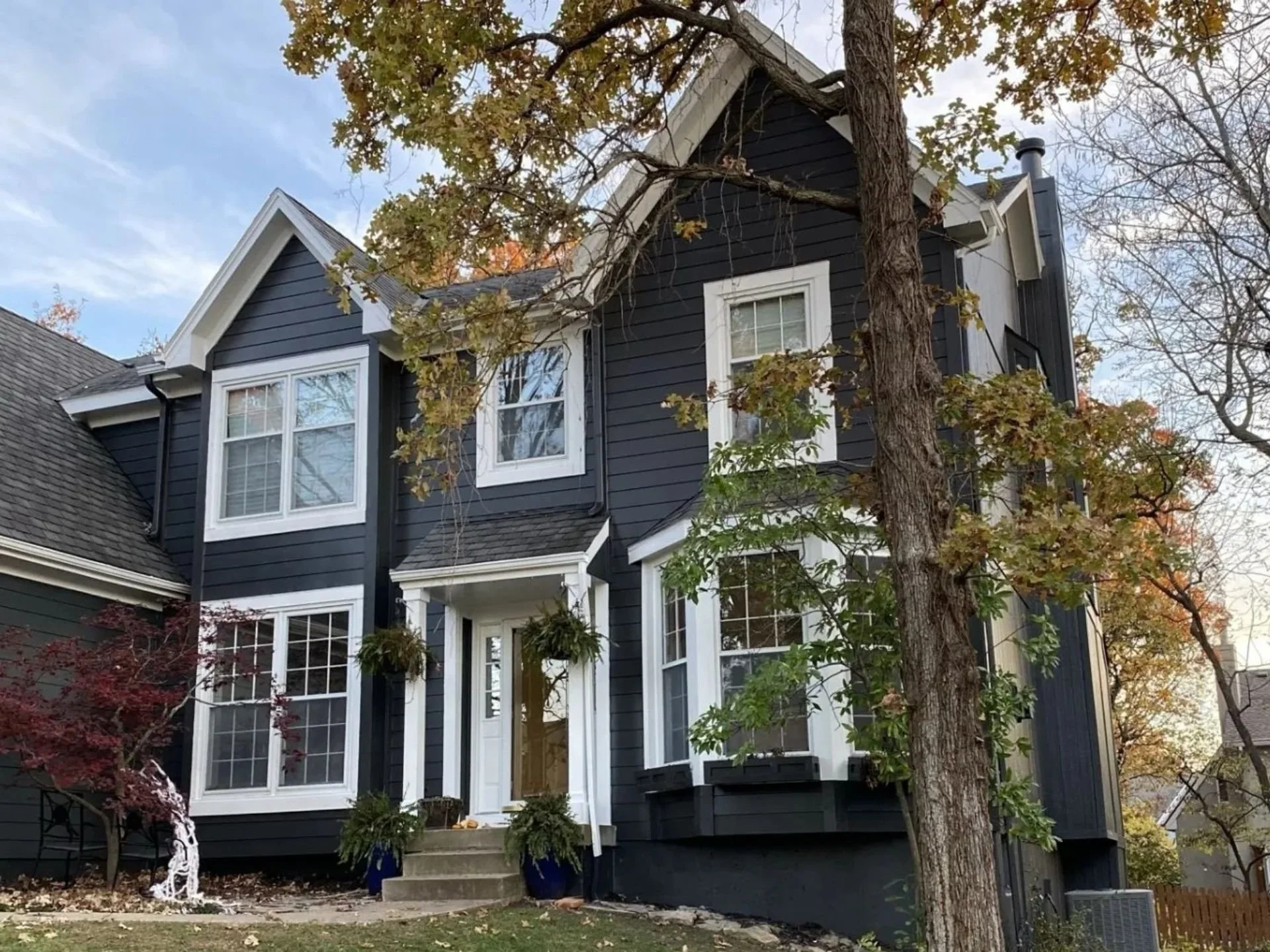 A newly painted dark navy blue multi-story house in Shawnee, Kansas with white trim and large windows, surrounded by trees with autumn-colored leaves. 

