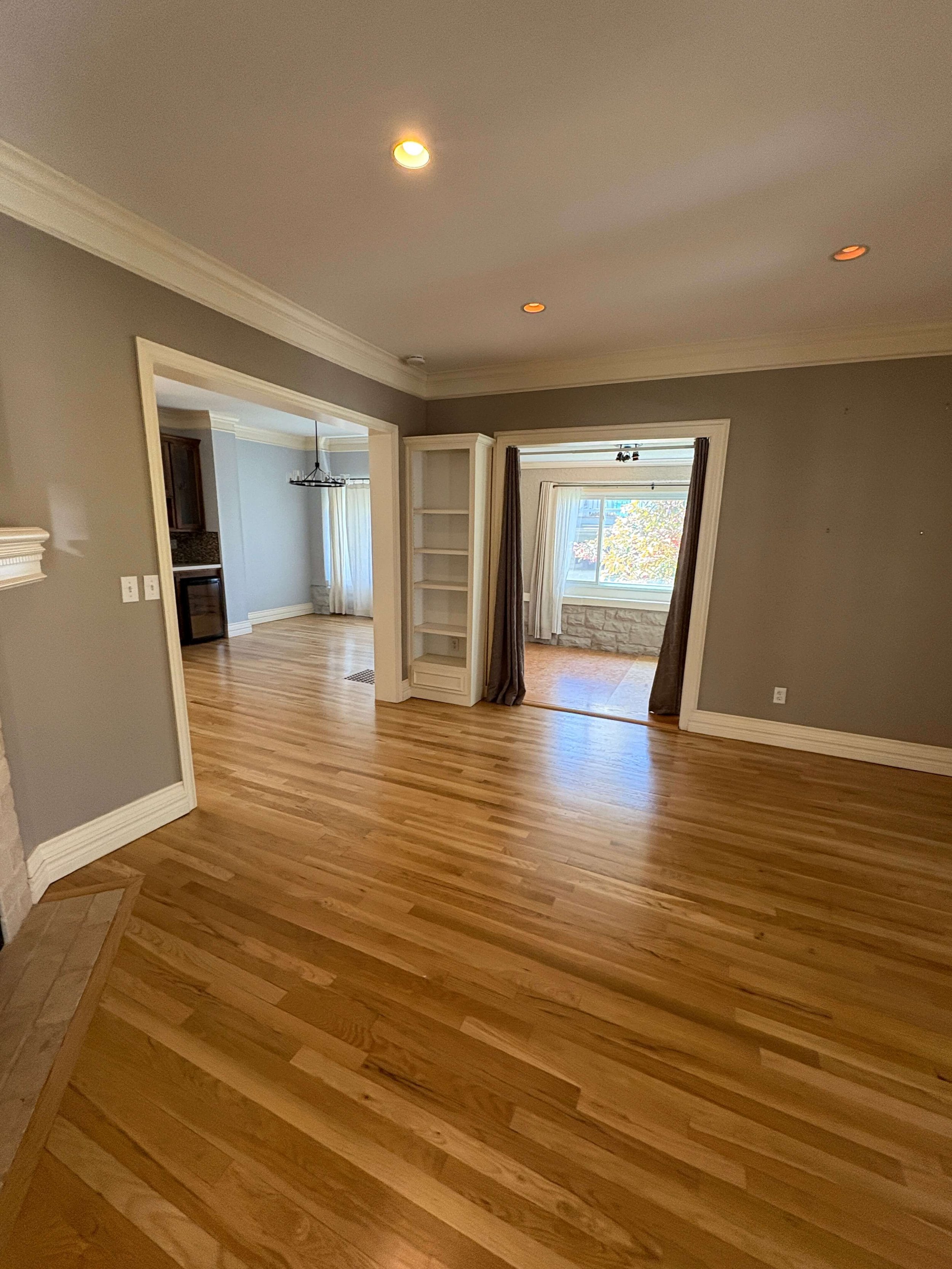View of a newly painted beige interior living space with wood flooring, a doorway leading to another room, and a window with curtains showing trees outside.
