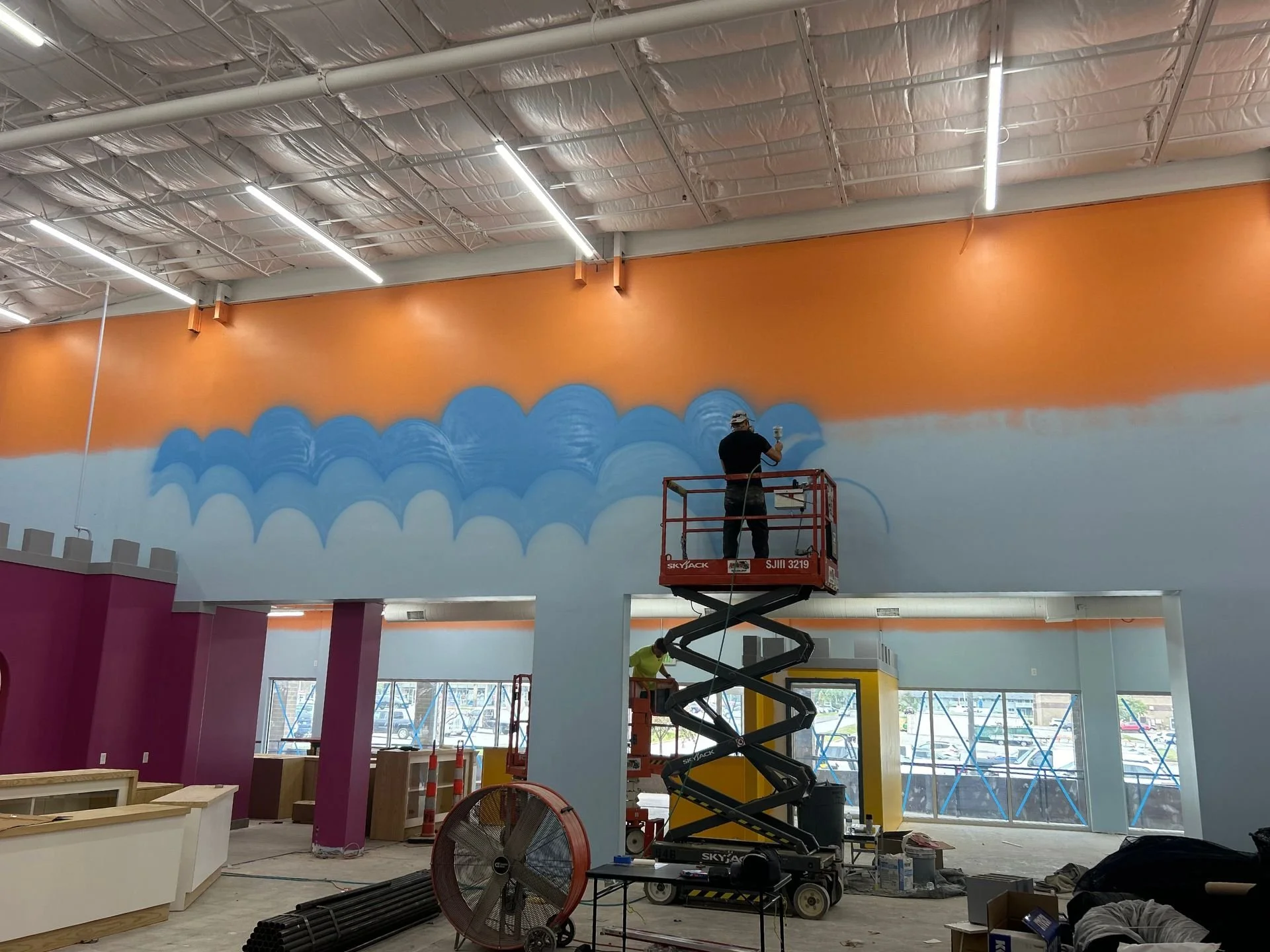 The interior of Kids Empire in Kansas while under construction. An LAK Painting painter is on a lift painting swirling blue clouds on a light-blue and orange concrete wall. There are purple walls and pillars and lighting above.