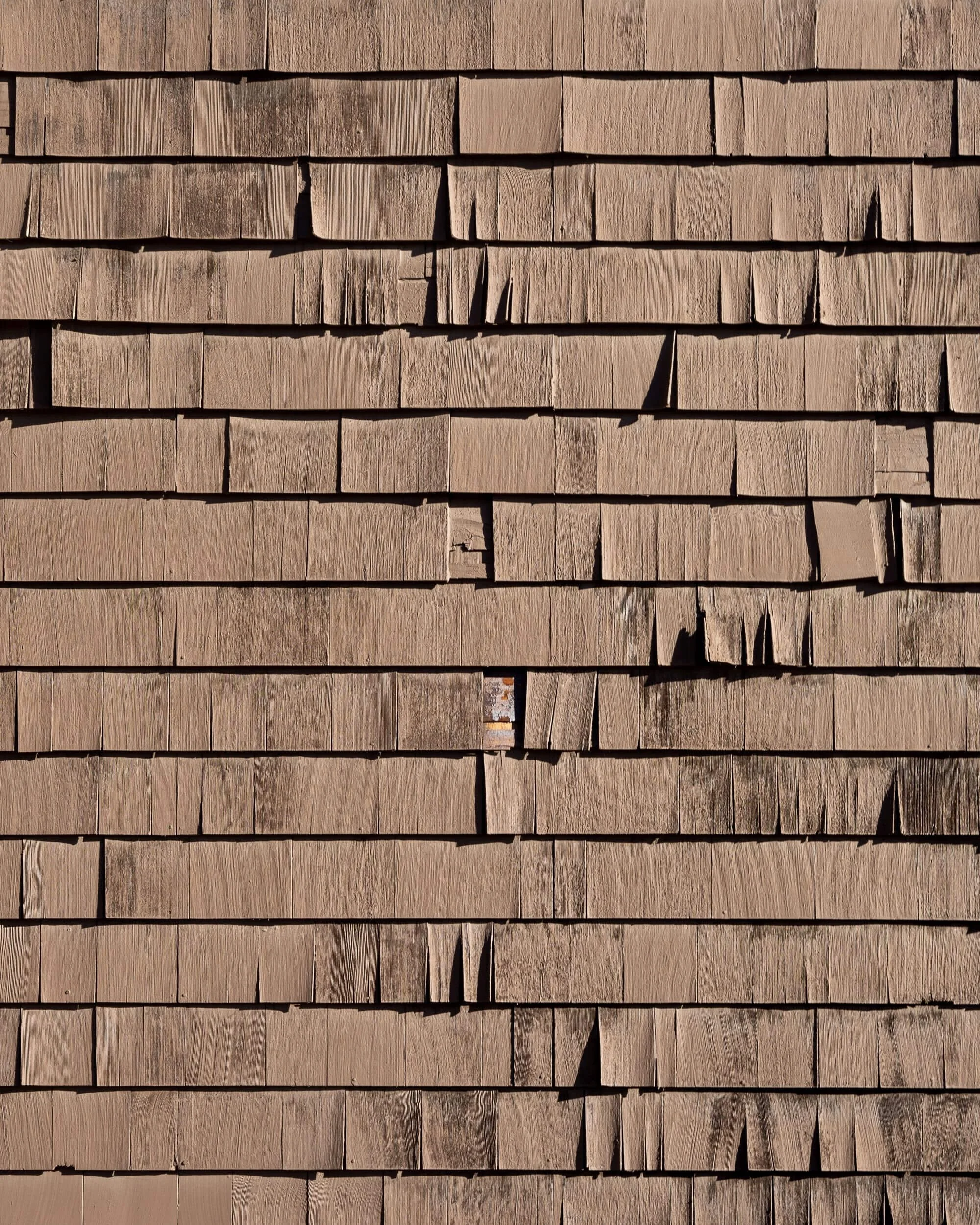 Close-up of a wooden shingle wall, showing weathered, uneven, and cracked shingles in shades of brown with wood rot in need of replacement.
