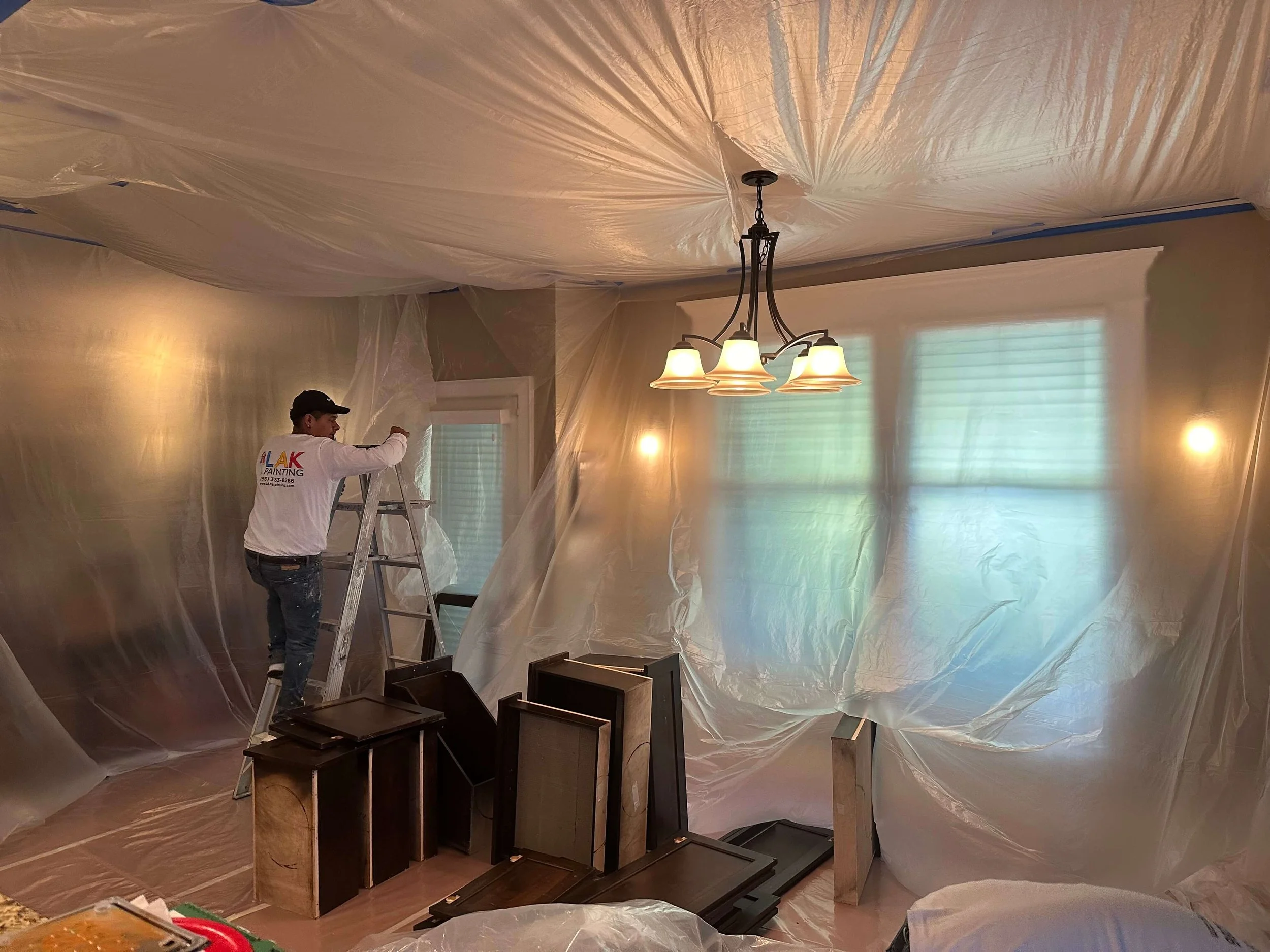 An LAK Painting painter with an LAK Painting t-shirt and jeans standing on a ladder taping plastic protective sheeting on the walls, windows and ceiling. There are cabinets to be painted on the floor.