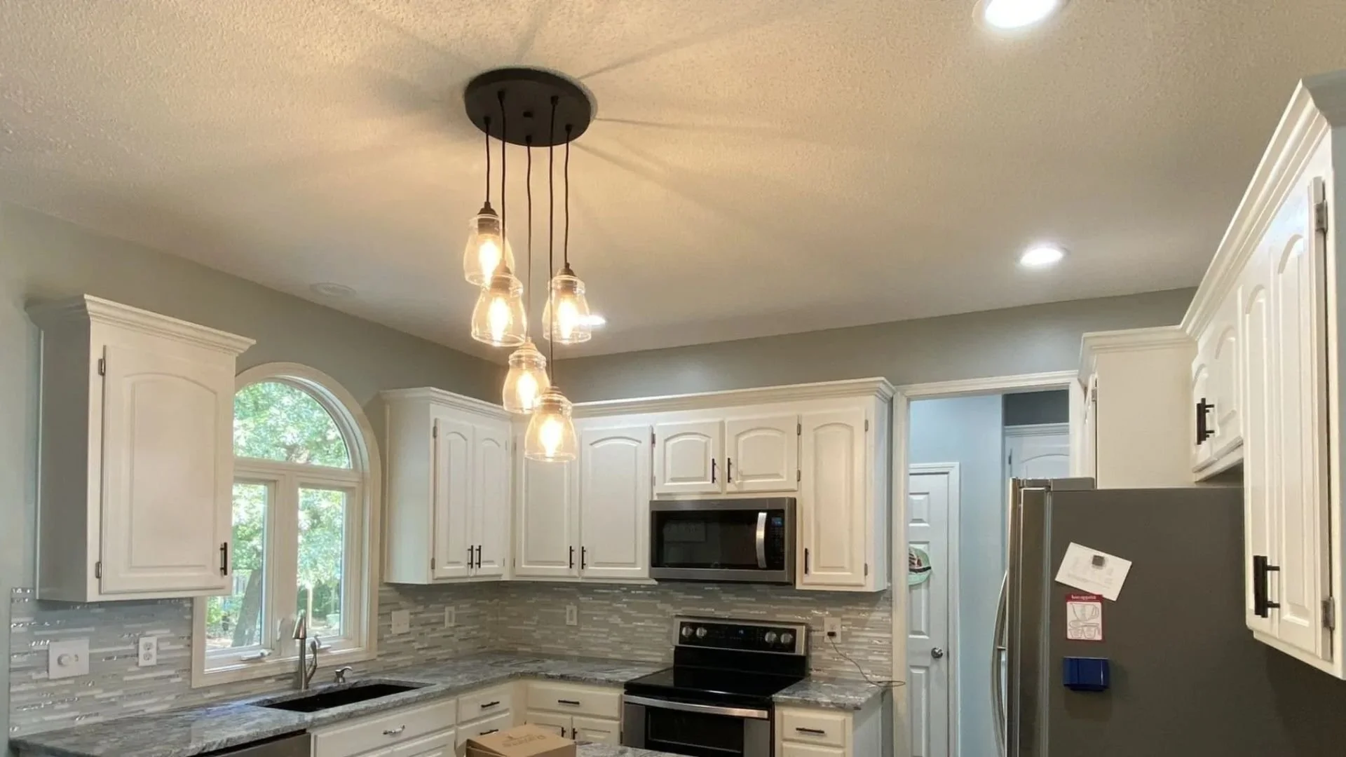 Kitchen with newly painted white cabinets, gray walls, a window, a microwave, stove, granite countertop, and a modern light fixture with hanging bulbs on the ceiling.
