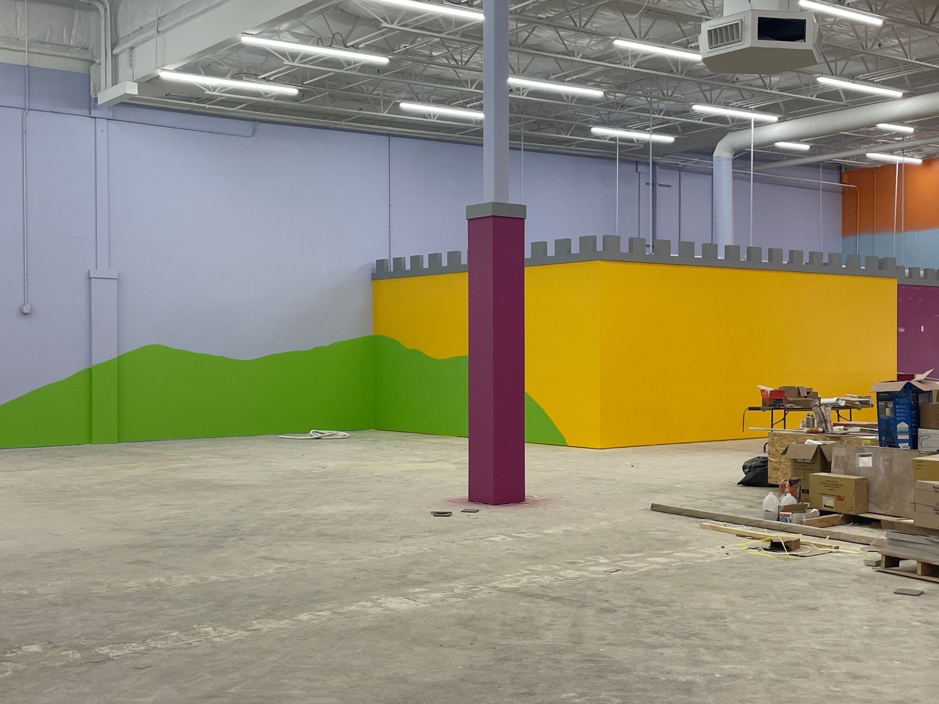 Interior of Kids Empire in Kansas with newly painted concrete walls, bare concrete floors and industrial lighting. The farthest wall is light blue with a yellow walled area on the right with a green hill painted motif going across both walls.