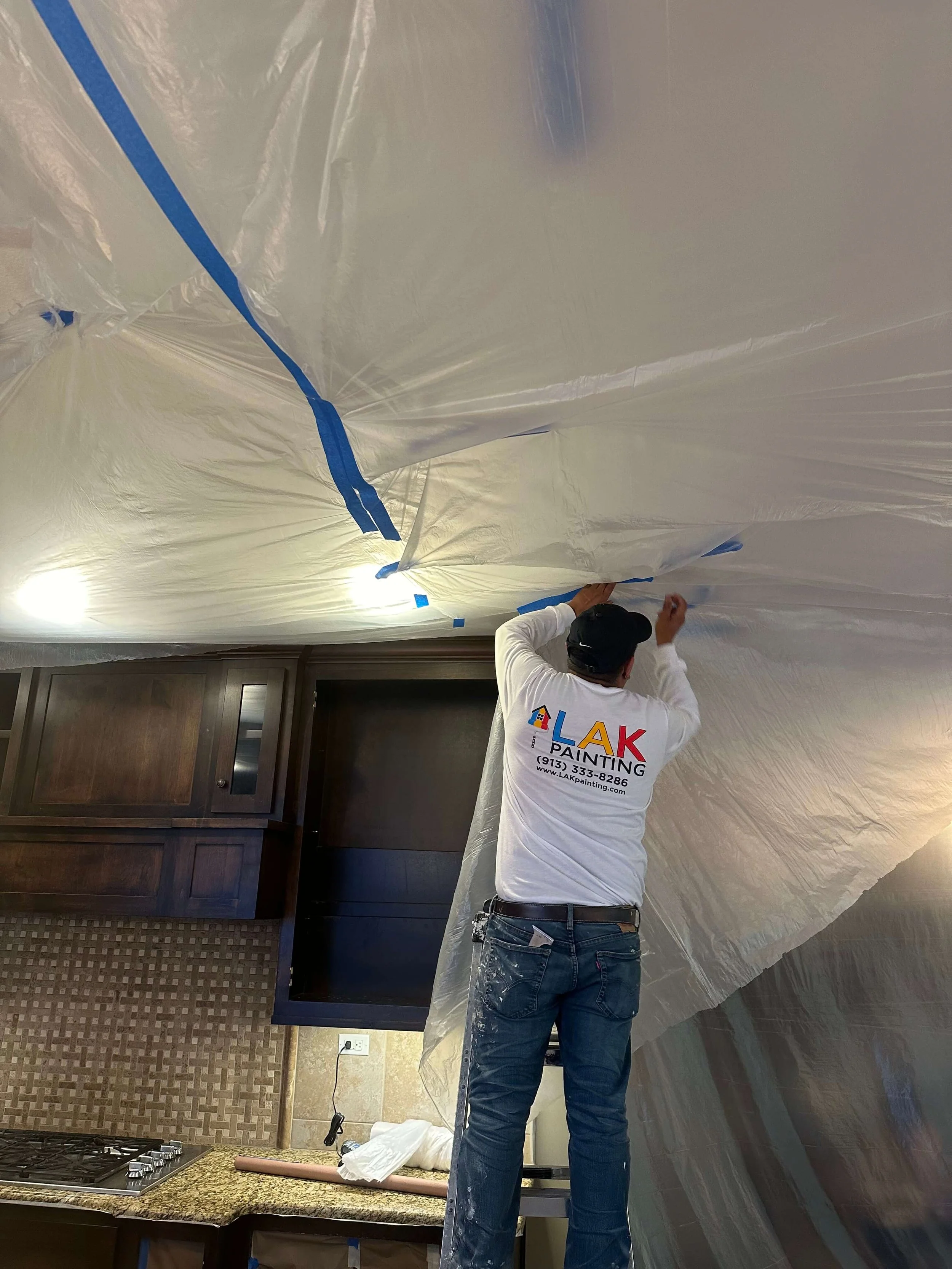 An LAK Painting painter with an LAK Painting t-shirt and jeans taping plastic protective covering on the ceiling of a furnished kitchen in a Lenexa, Kansas home.