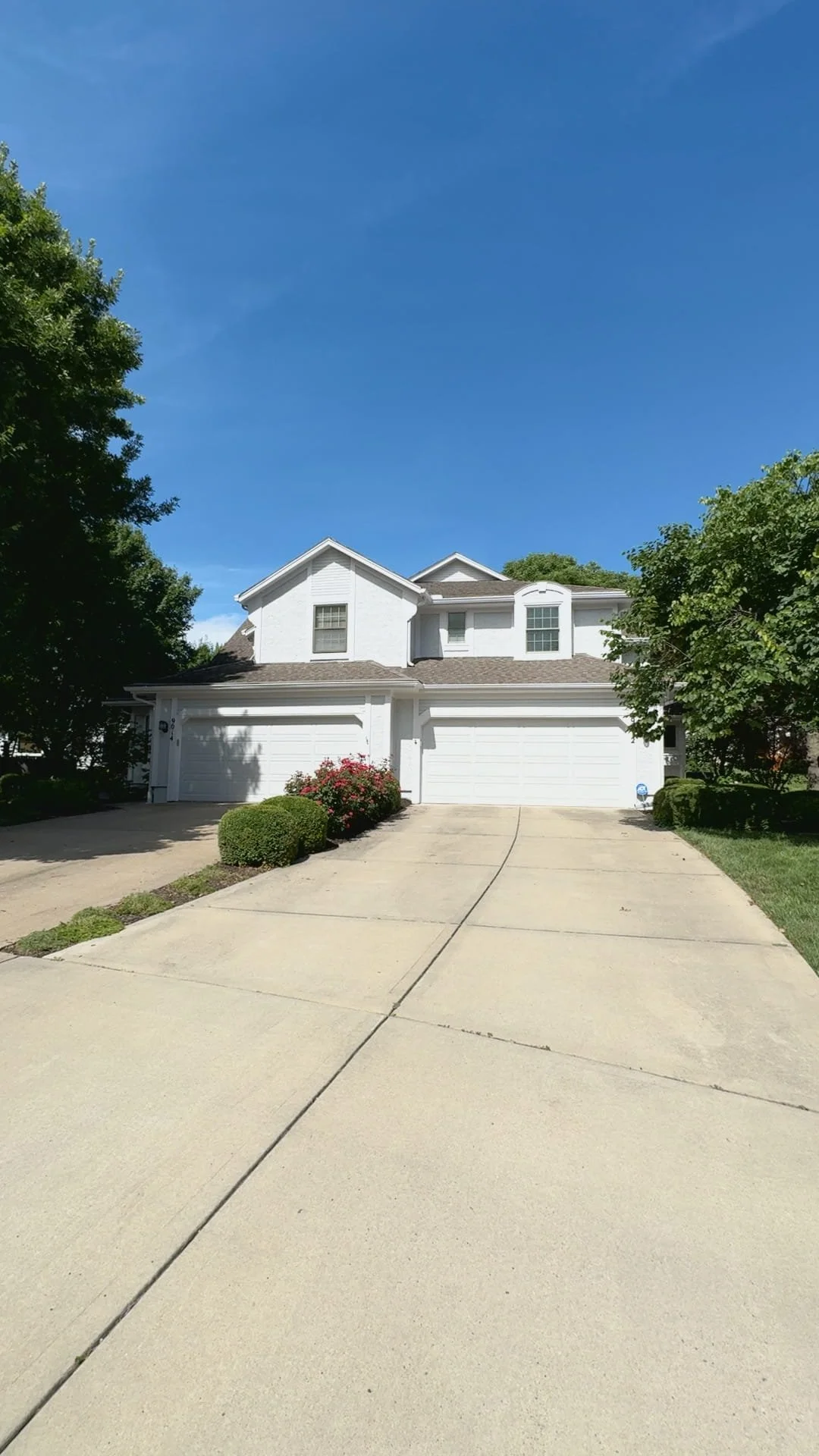 A white, two-story house with a three-car garage, surrounded by green trees and bushes, under a clear blue sky.