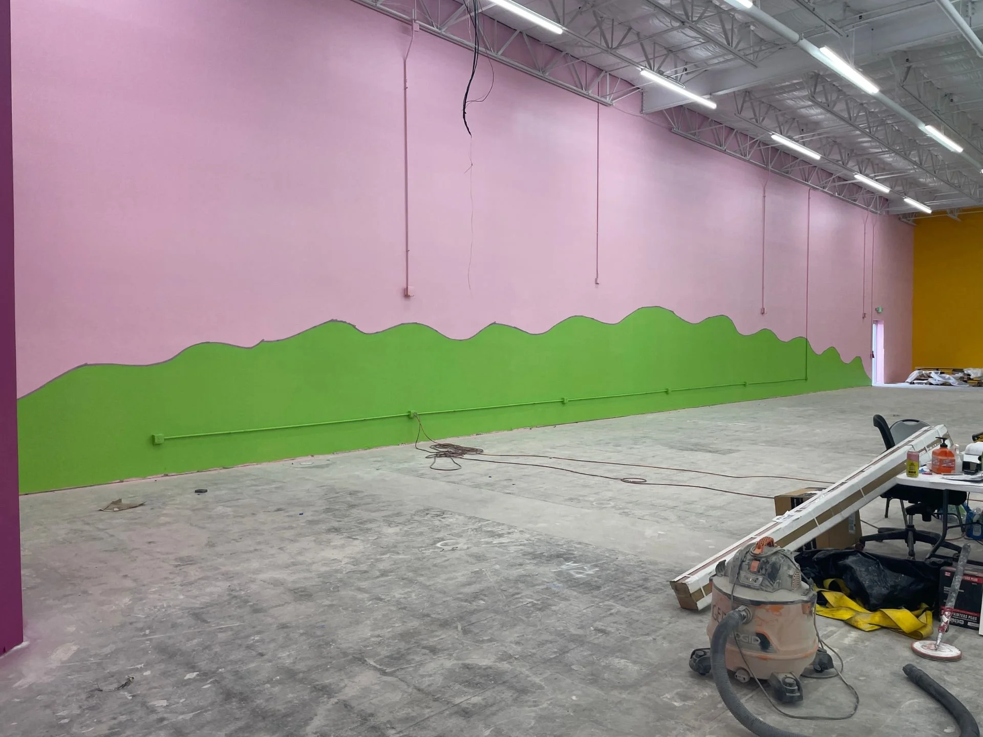 Interior wall of Kids Empire in Kansas with newly painted concrete walls, bare concrete floors and industrial lighting. The wall is pink with a green hill painted motif going across both walls. There are construction materials on the floor.