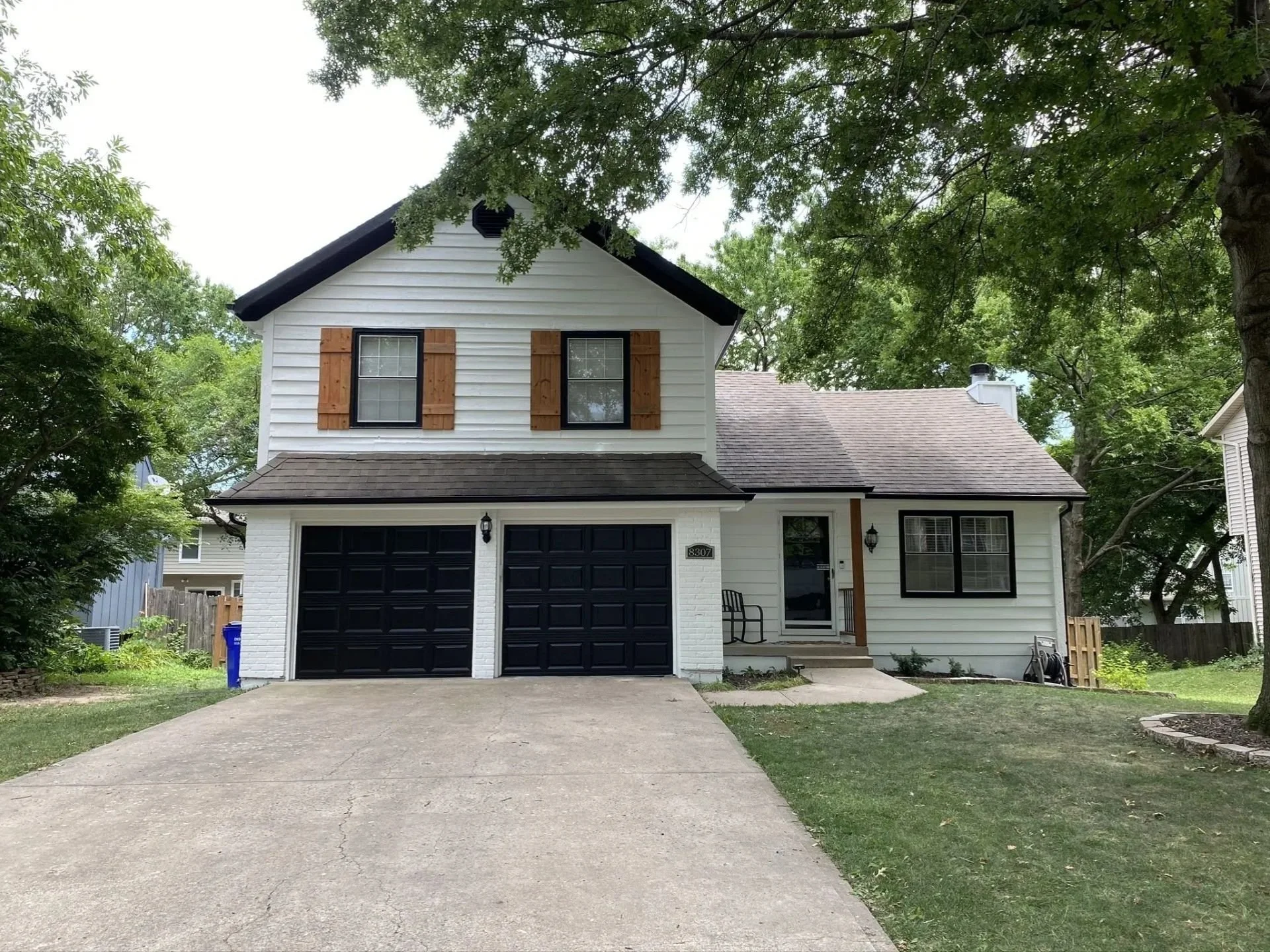 White two-story house in Shawnee, Kansas with black garage doors, wooden window shutters, and a small front porch, surrounded by trees and a well-maintained lawn.
