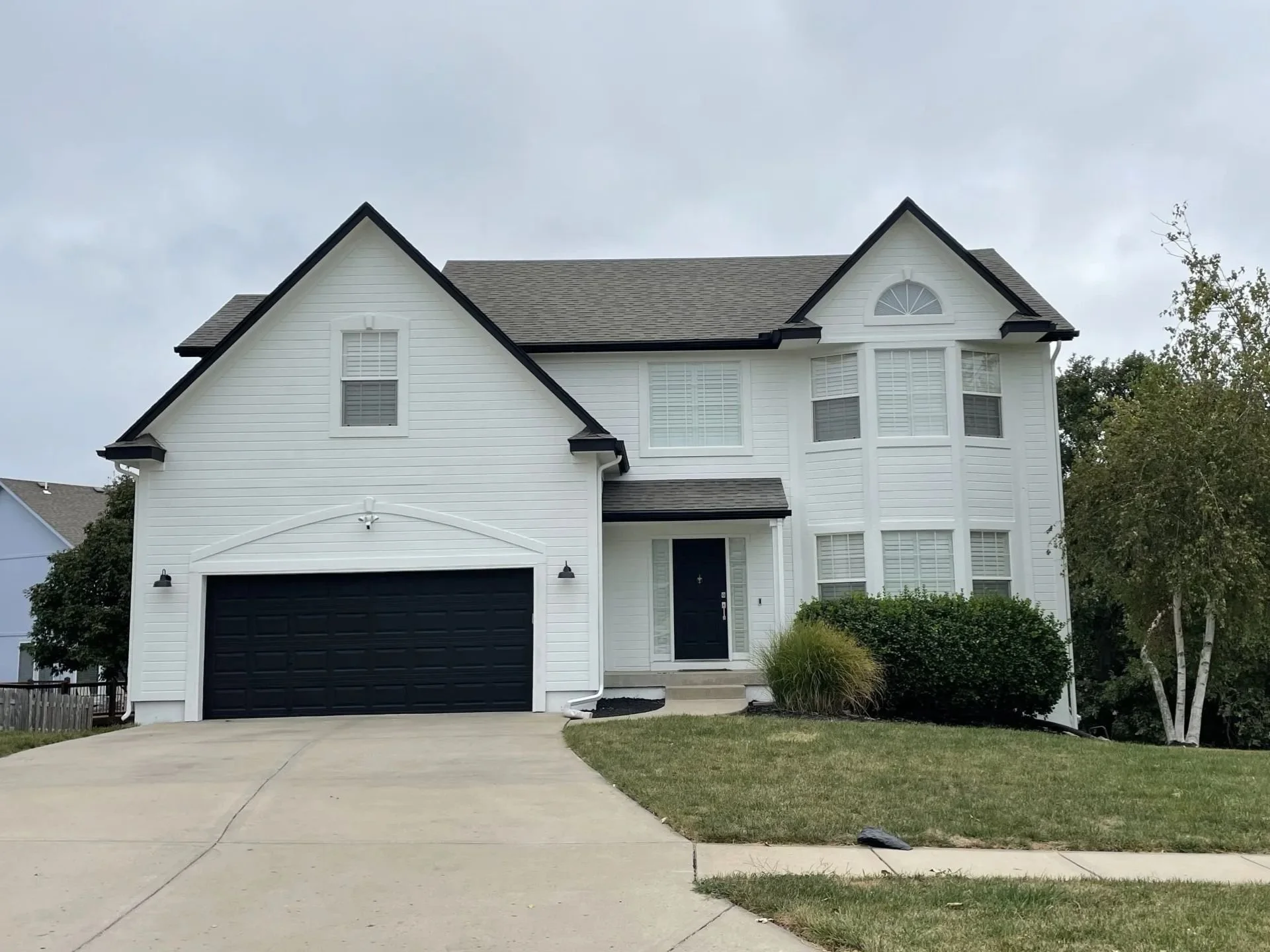 A newly painted white two-story house in Kansas City with a black garage door, black trim and windows with white shutters. A concrete driveway and sidewalk with a lawn with bushes.
