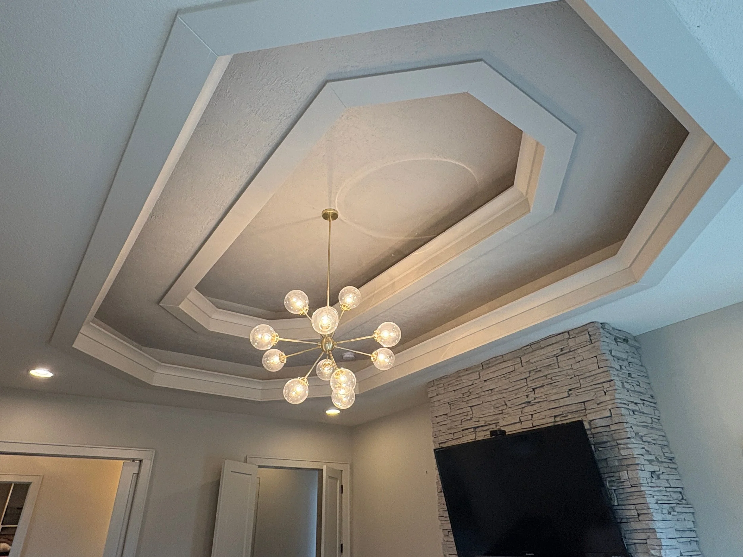 Ceiling view of a room with a newly painted wood oval inset, a modern chandelier with multiple globe-shaped lights, and a stone fireplace below the ceiling.