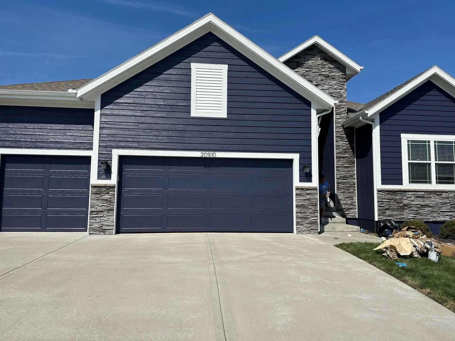Modern two-story house in Johnson County, Kansas with newly painted navy-blue horizontal siding, stone accents, a two-car garage, and a front porch. The sky is blue.