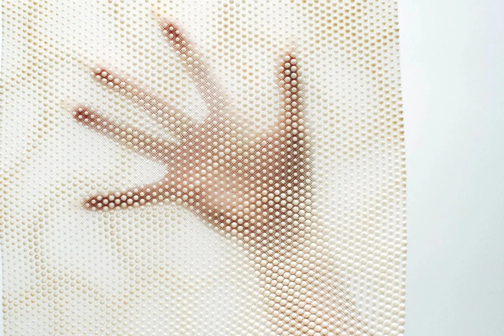 A hand holding a square tactile surface panel covered in dense white raised dots, with its silhouette visible through the texture