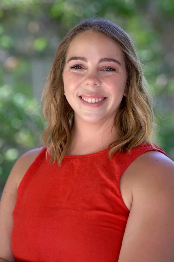 A young woman smiling outdoors, wearing a red sleeveless top, with a background of green blurred trees.