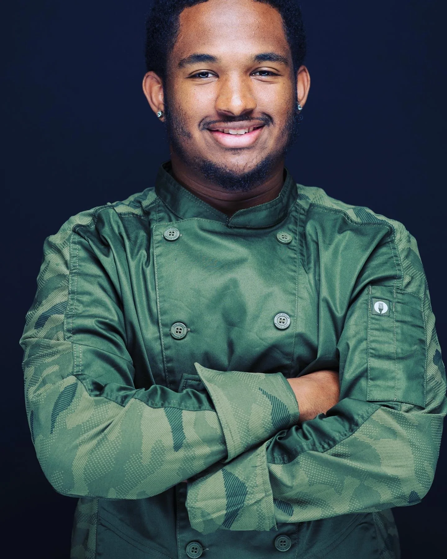 Portrait of a young man with short curly hair, earrings, and a well-groomed beard, smiling with arms crossed, wearing a green chef’s jacket, against a dark background.