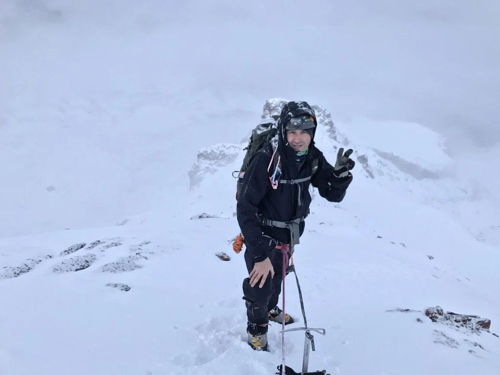 Chris Knuth posing at the top of a snow-covered mountain, holding up a peace sign