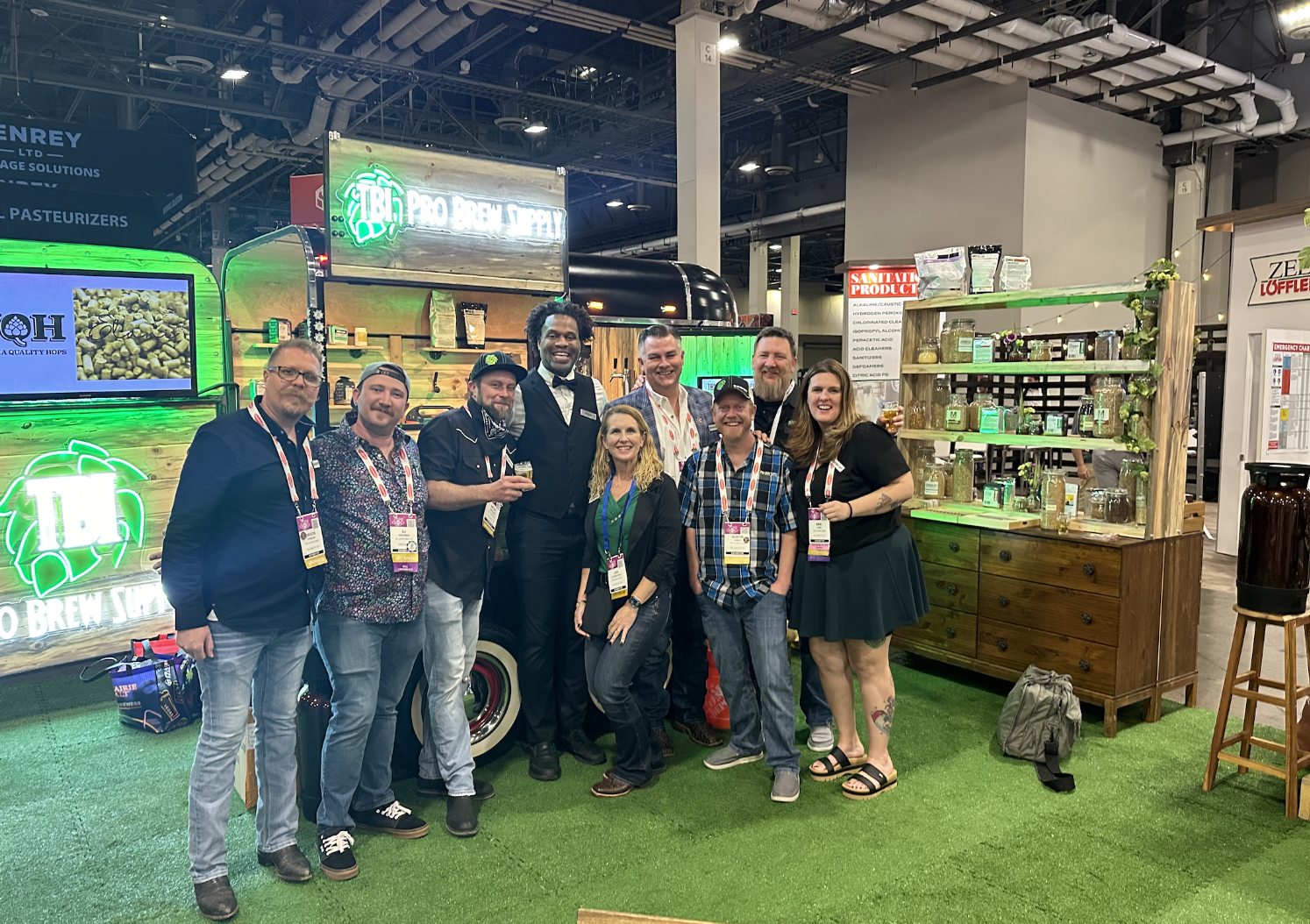 Group of people standing in front of a booth at a convention or trade show, with beer brewing and health product displays, some holding drinks, smiling for the camera.