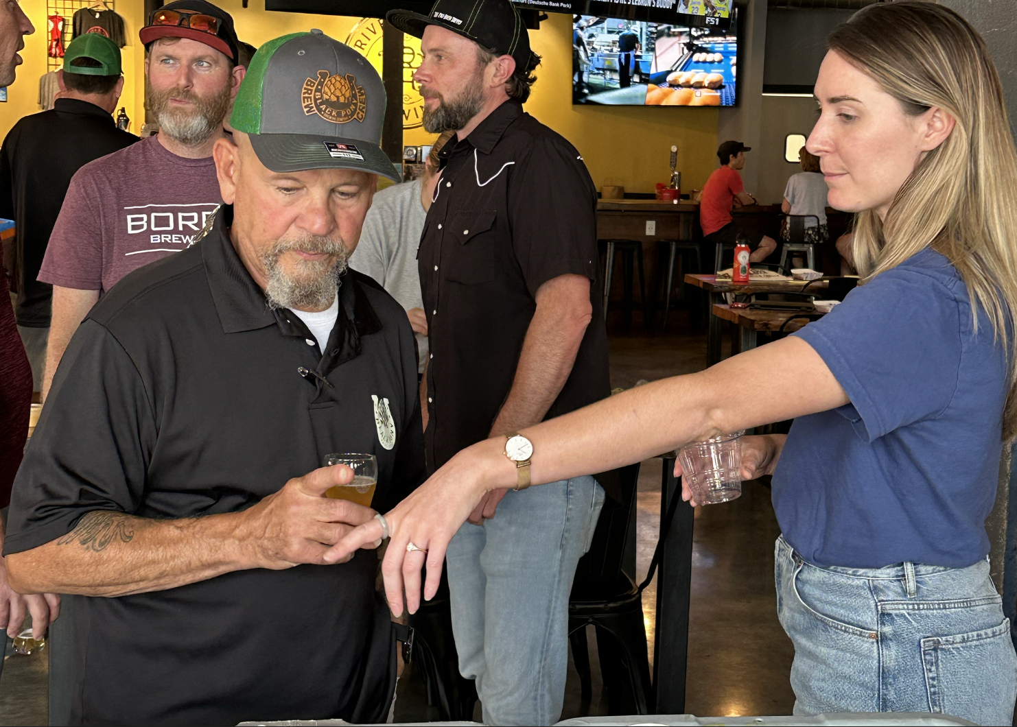 A young woman with blonde hair in a blue shirt is speaking to to an older man with a gray beard, wearing a black polo shirt and a baseball cap, inside a bar or restaurant with several other people in the background.