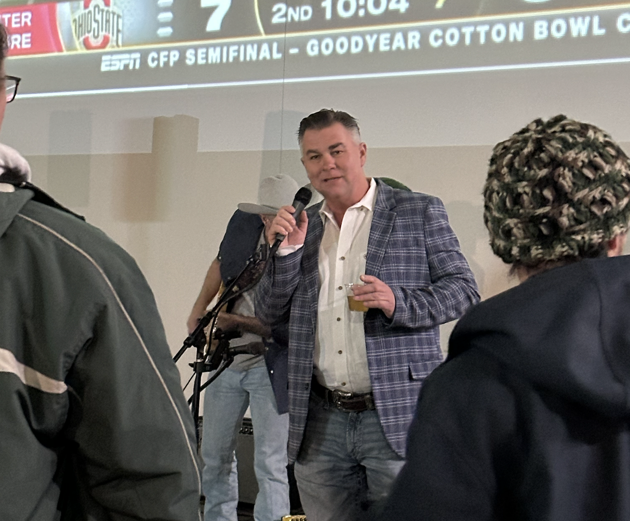 A man in a blue plaid blazer speaking into a microphone at an indoor event, holding a drink, with several people listening in the foreground