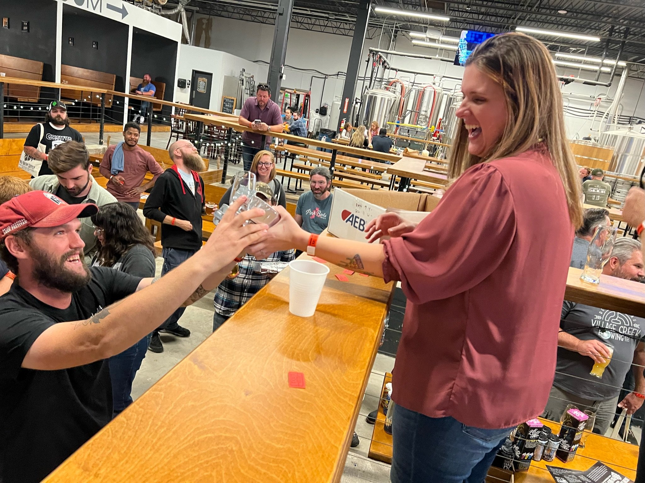 A group of people socializing at a brewery or taproom, with some sitting at tables and others standing. A woman in a pinkish-brown shirt is smiling and handing a glass to a man with a beard. The background features brewing equipment and more patrons.