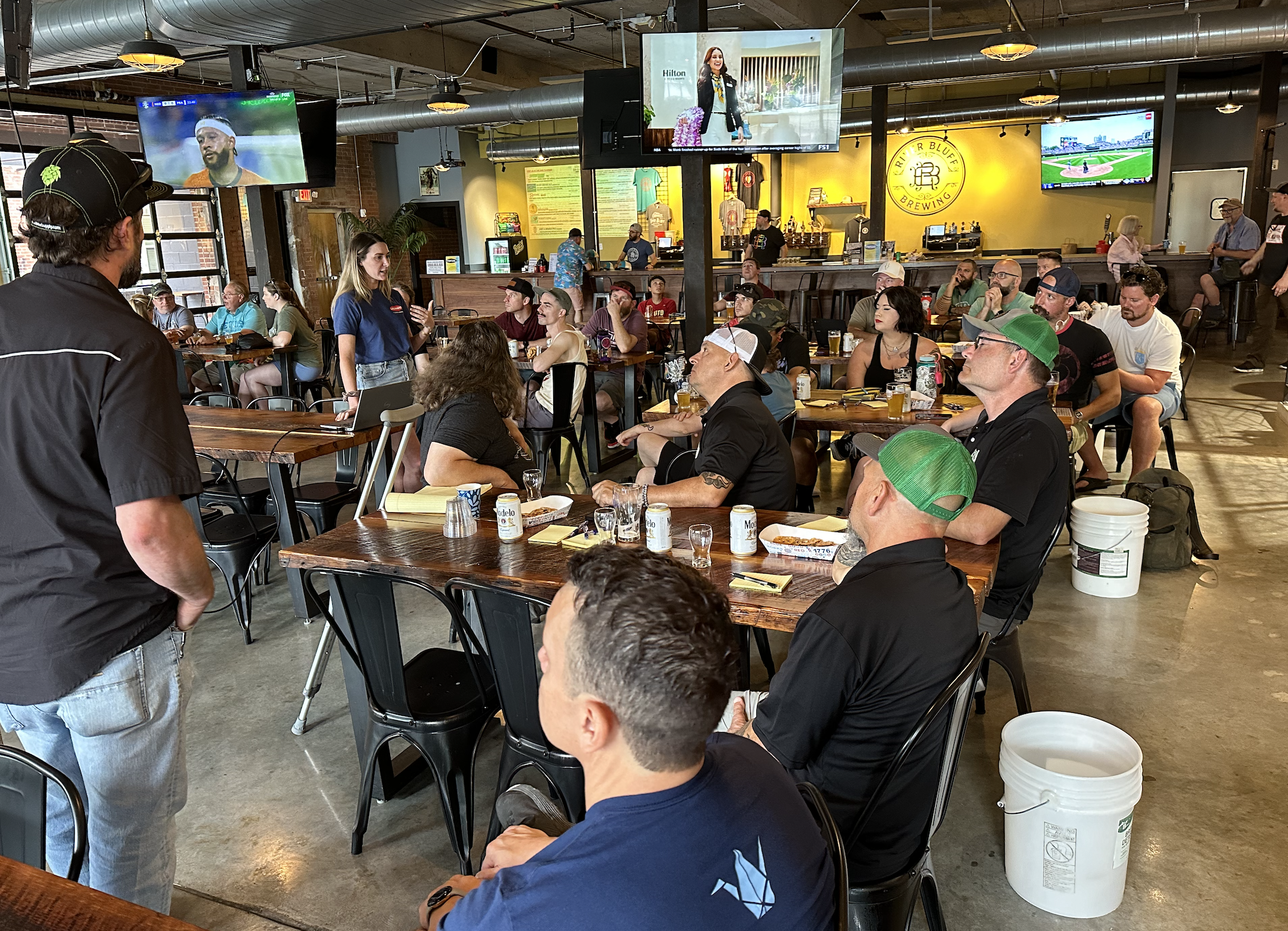 People gathered inside a brewery or bar, watching a presntation, with some sitting at tables drinking beer and others standing or walking around.