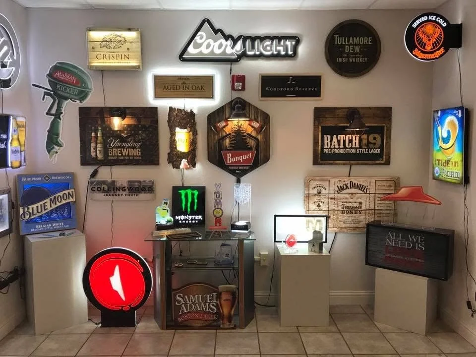 Collection of illuminated signs and decor on a wall, including beer brand signs, neon lights, and a small display table with electronic devices and small items.