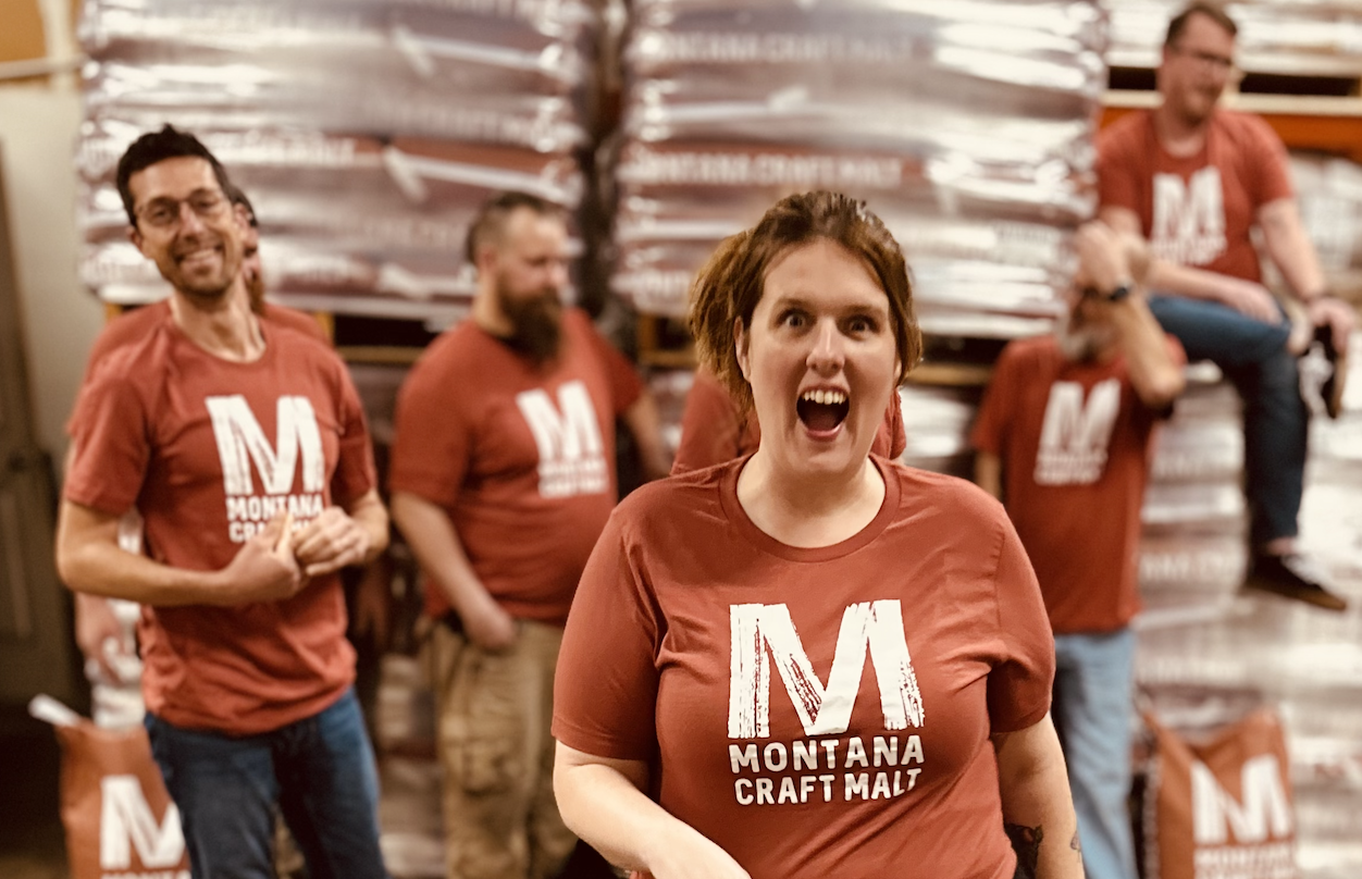 Group of people wearing red Montana Craft Malt T-shirts, standing and sitting in front of stacks of malted grains at a brewery or craft malt facility.
