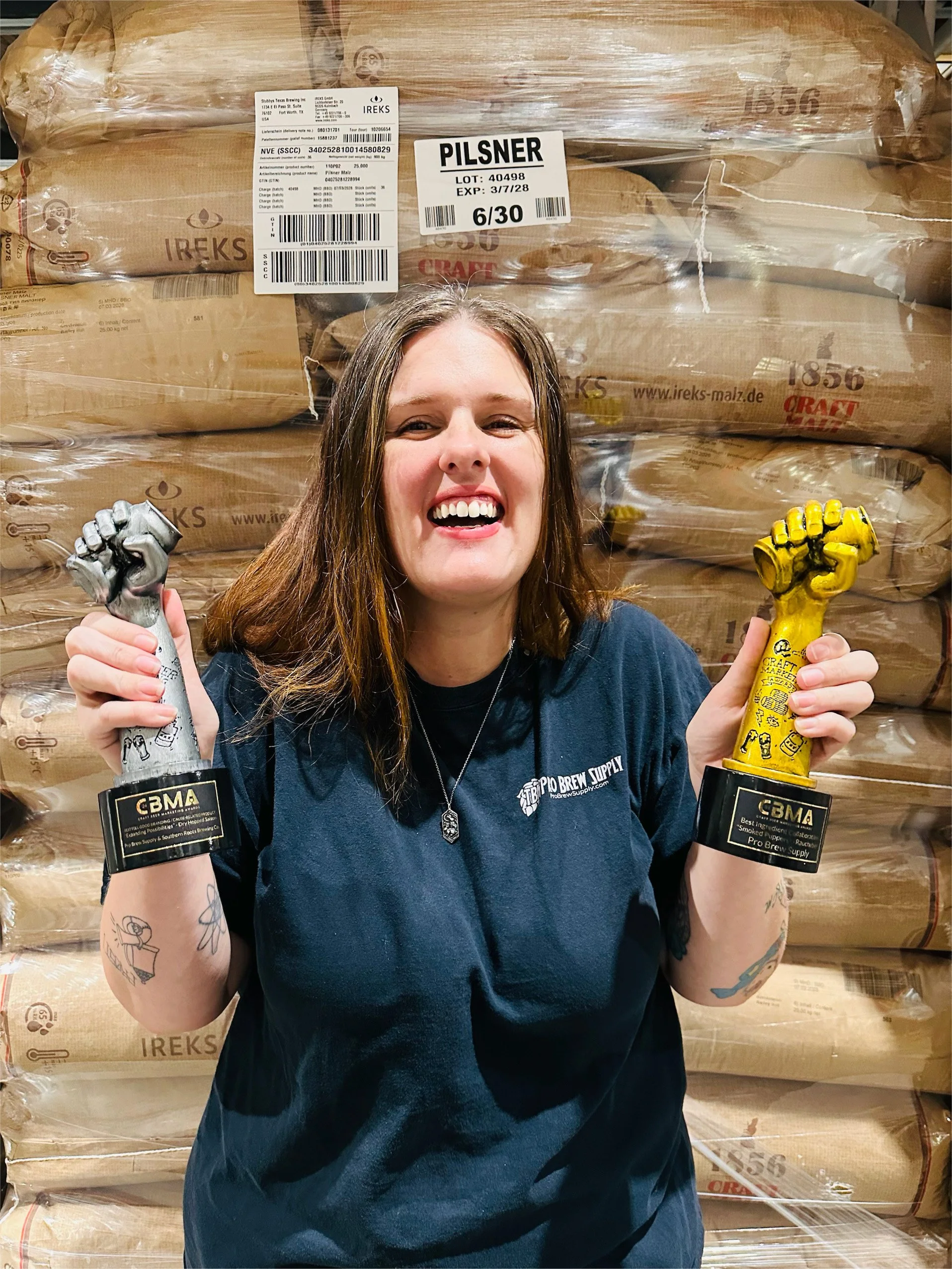 A smiling woman with tattoos on her arms, holding a silver fist-shaped trophy in her left hand and a gold fist-shaped trophy in her right hand, standing in front of stacks of large brown paper bags wrapped in plastic at a brewery or warehouse.
