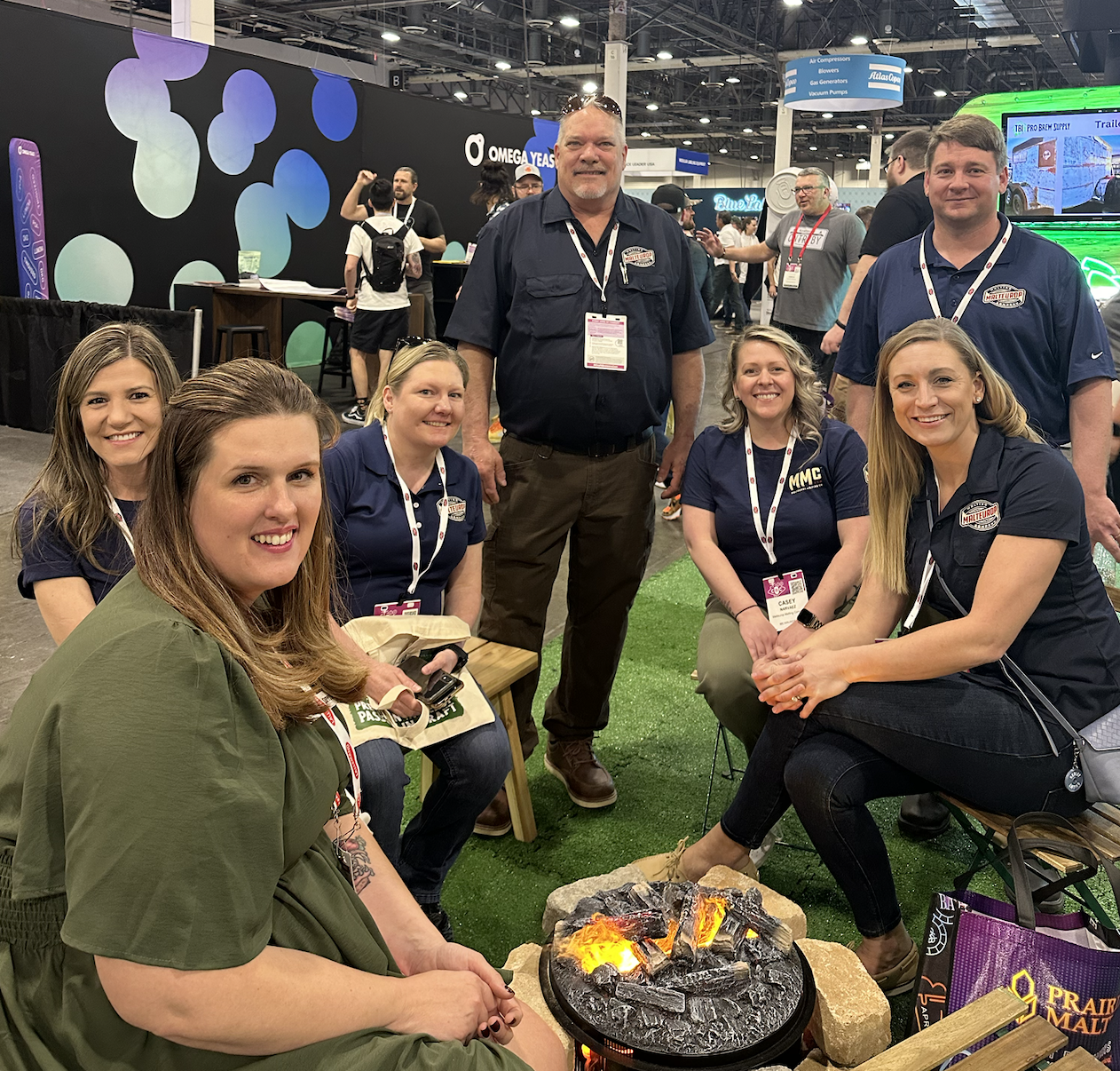 Group of six people at a conference or expo, sitting and standing around a small fire pit with glowing embers, in a busy indoor convention hall with signage in the background.